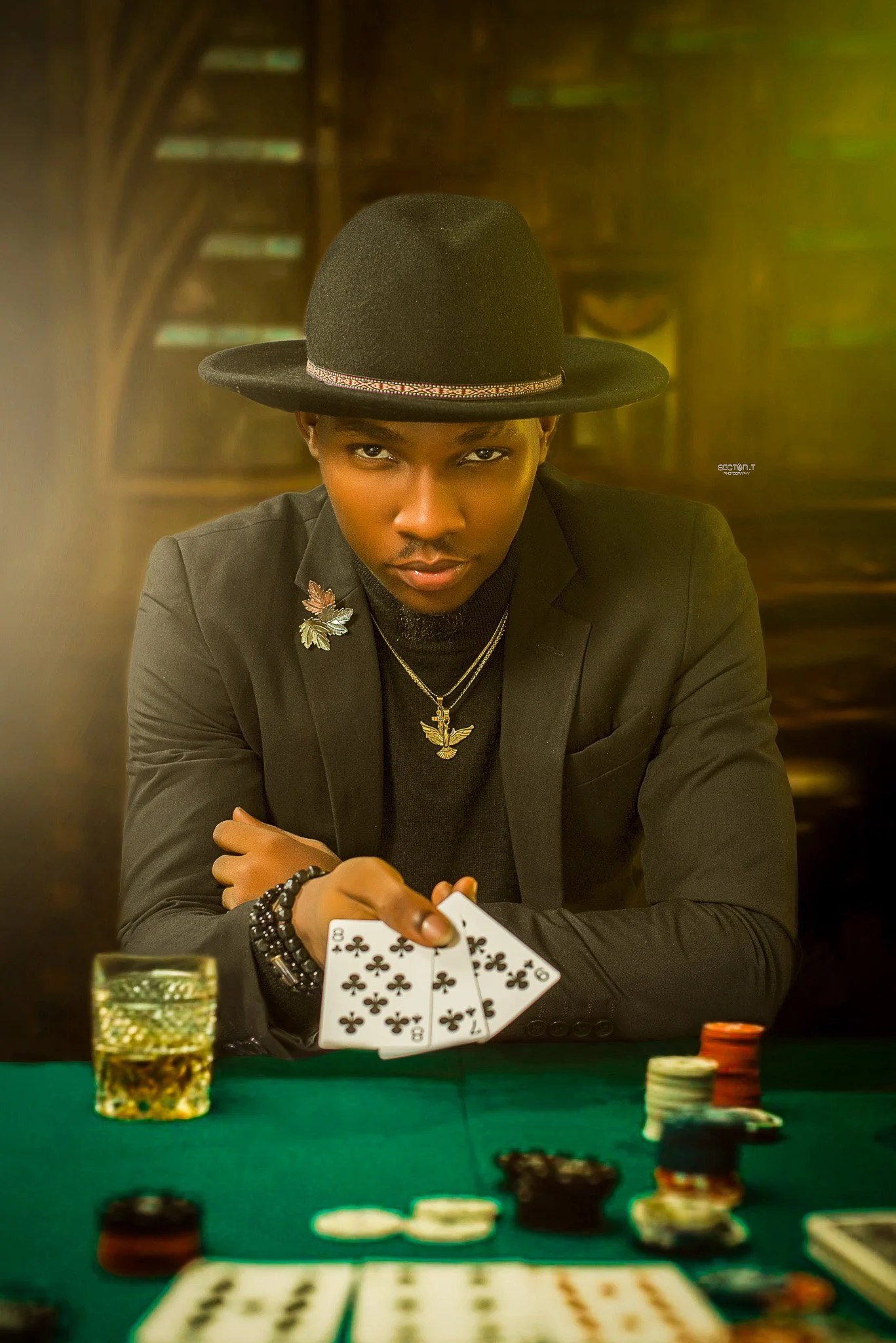 A young man dressed in black with a wide-brimmed hat, holding playing cards, sitting at a poker table with poker chips and a glass of beverage, in a dimly lit environment.