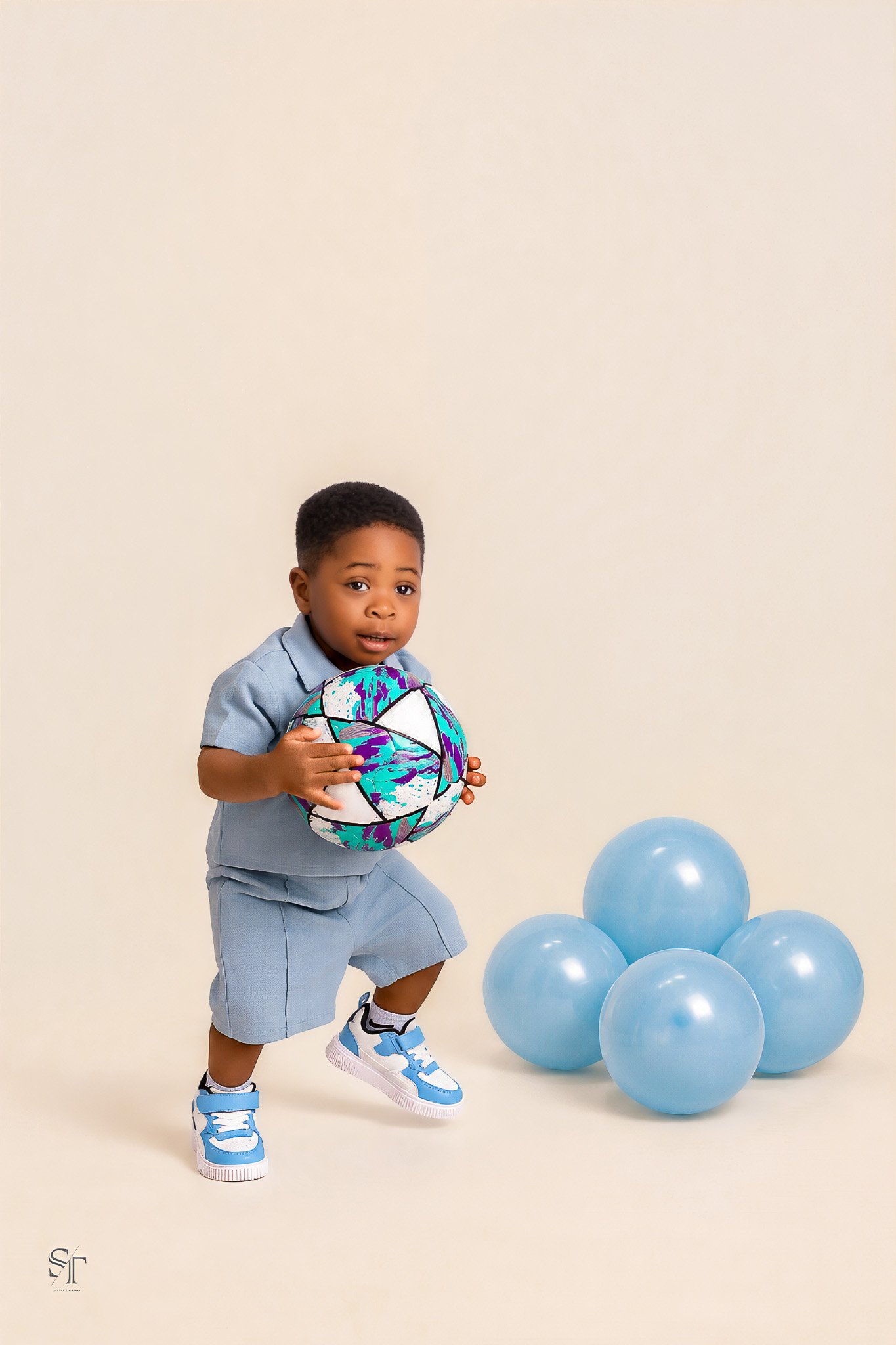 Young boy dressed in a light blue outfit holding a colorful soccer ball, surrounded by blue balloons against a plain beige background.