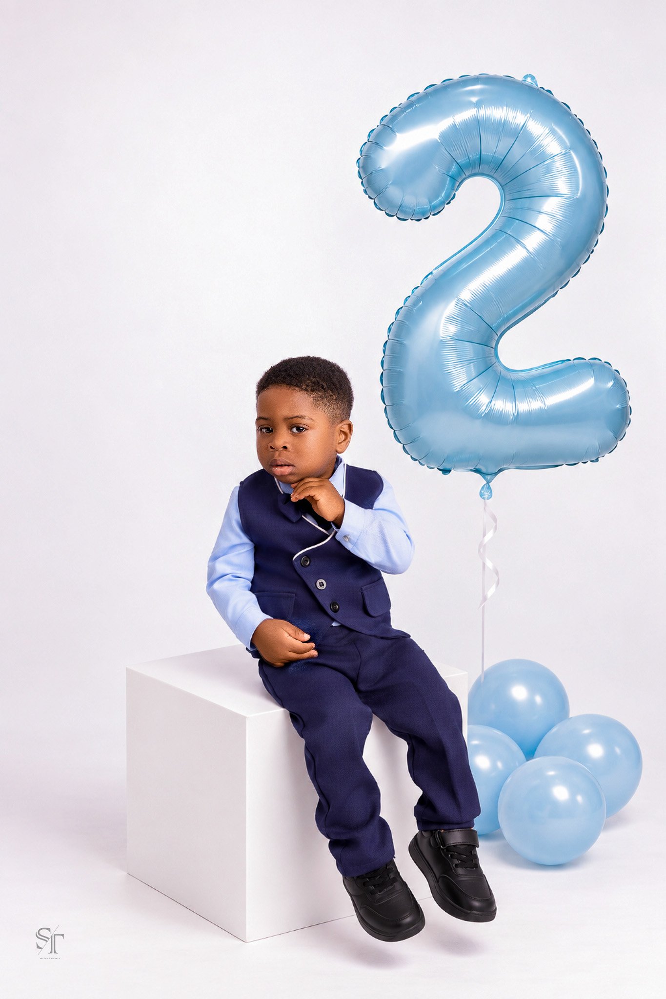 Young boy in navy blue suit sitting on a white block, with a large light blue number 2 balloon and several light blue balloons nearby, against a plain white background.