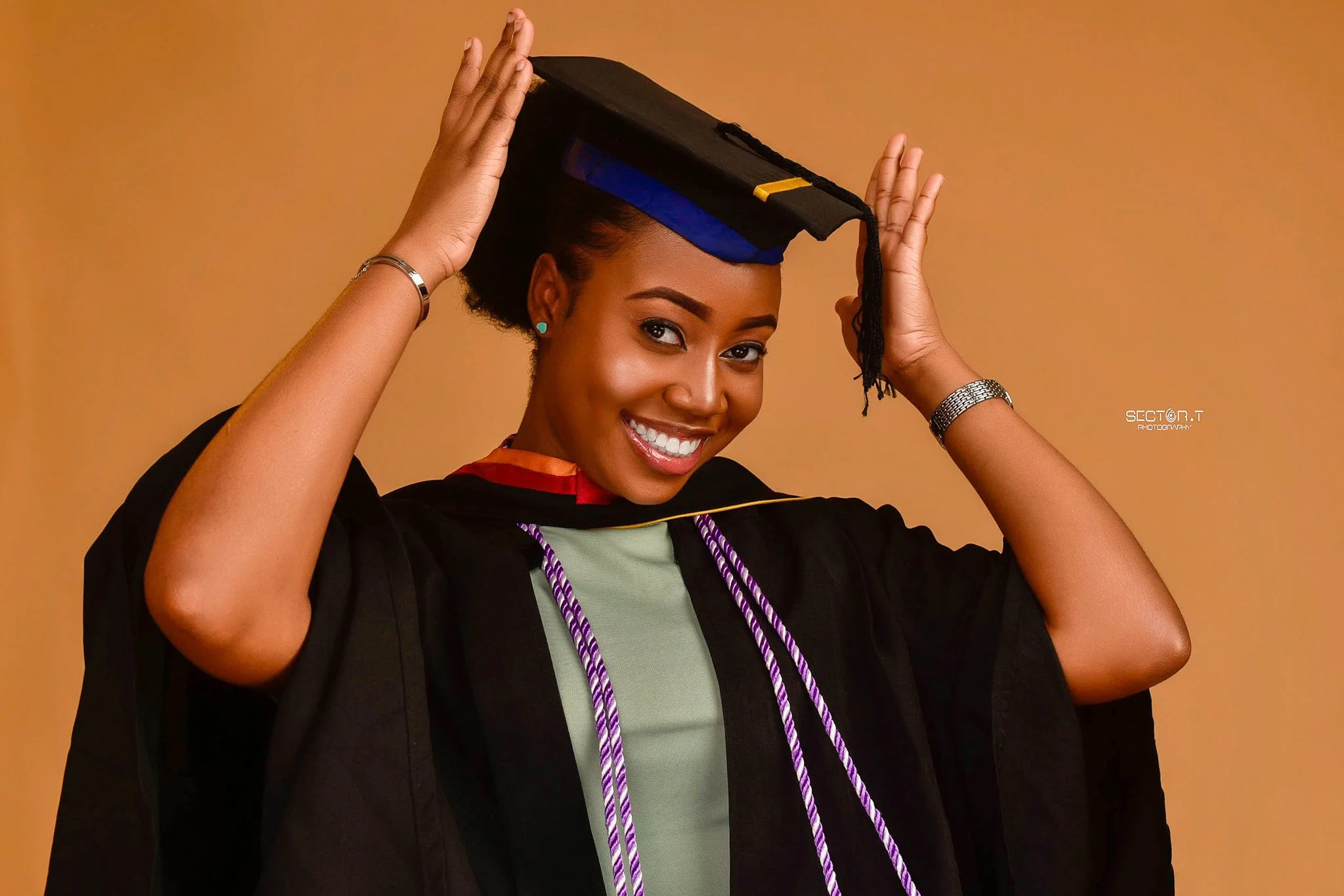 Happy young woman in graduation cap and gown adjusting her cap against a plain background.