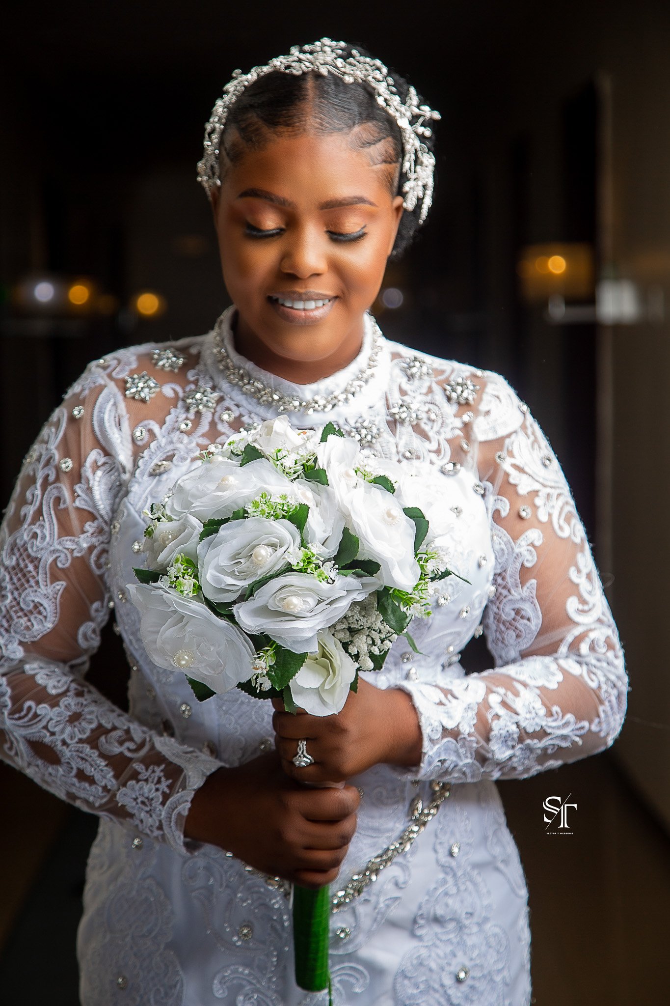 A bride wearing a white wedding dress with lace and pearl embellishments, holding a bouquet of white flowers, and adorned with a pearl headpiece and jewelry.