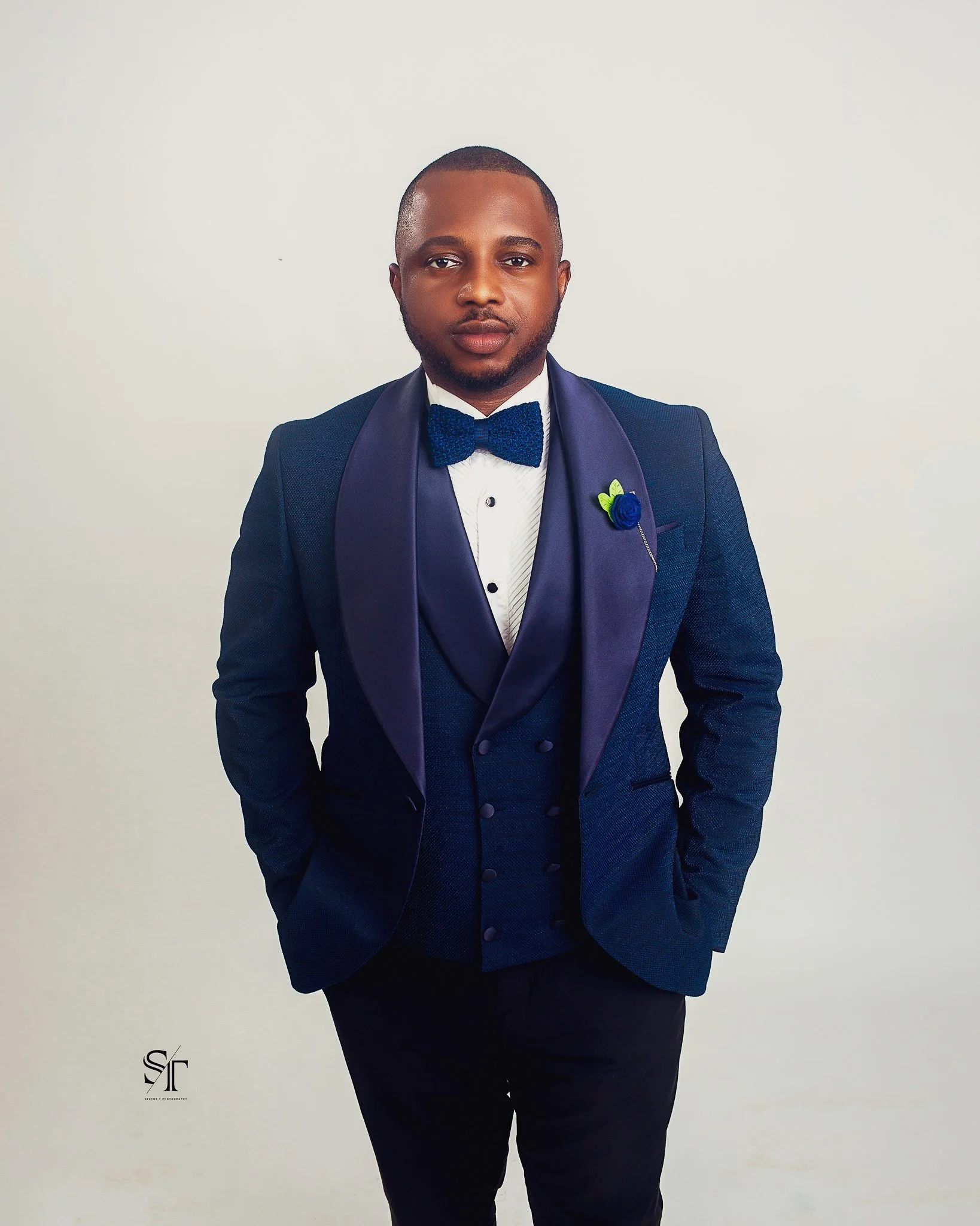 Man in a dark blue tuxedo with a satin shawl lapel, white shirt, dark blue bow tie, and a flower lapel pin, standing against a plain white wall.