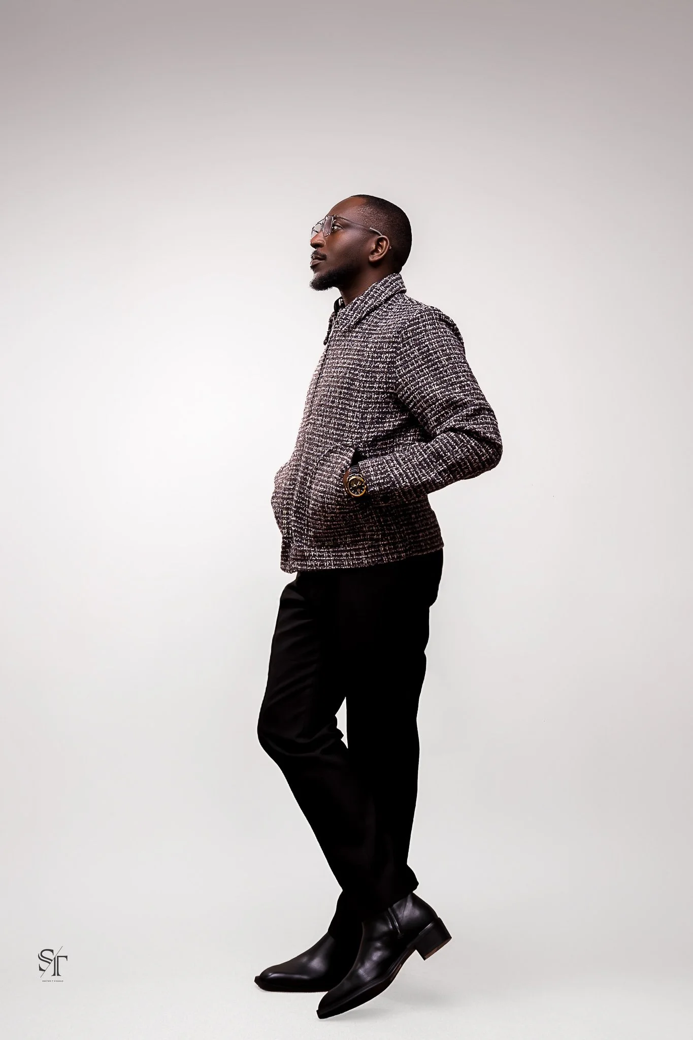 A man standing in profile with his hands in his pockets, wearing glasses, a patterned blazer, black pants, and black boots, against a plain white background.