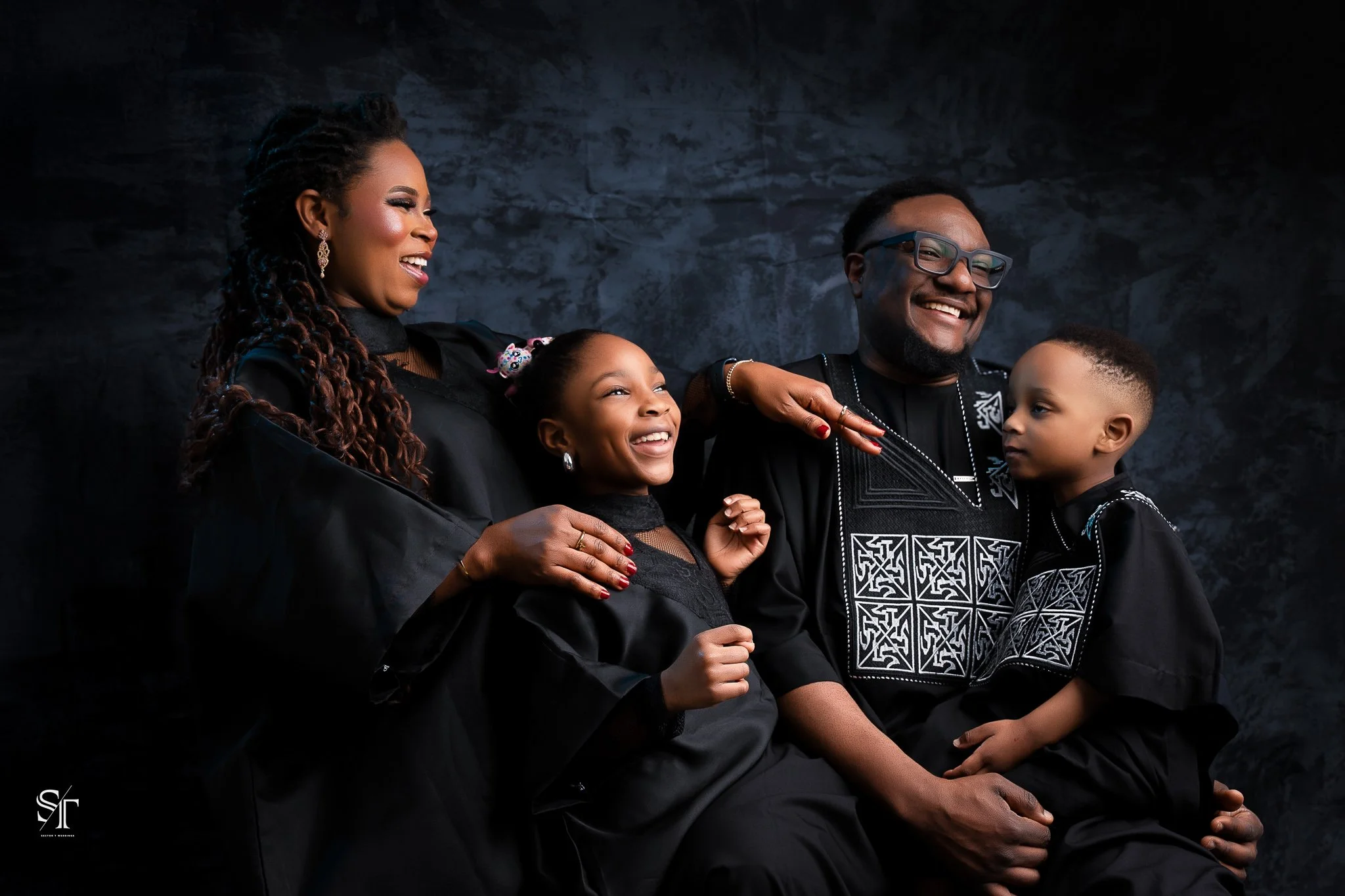A family of four with African descent dressed in black traditional clothing, smiling and laughing together against a dark textured background.
