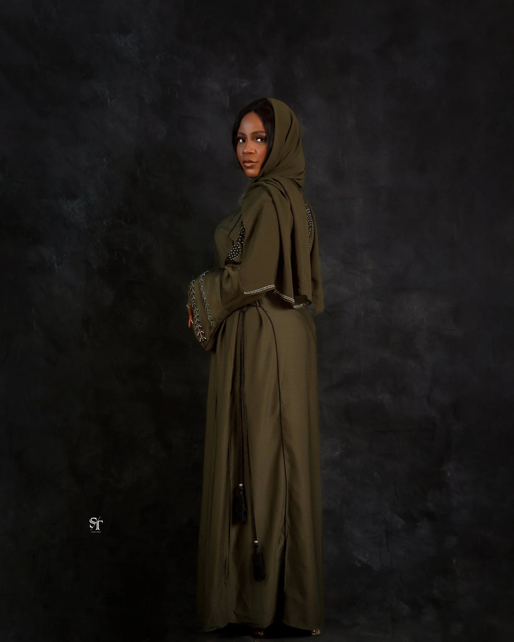 A woman in an olive green hijab and matching flowy dress, standing against a dark textured backdrop, looking over her shoulder.