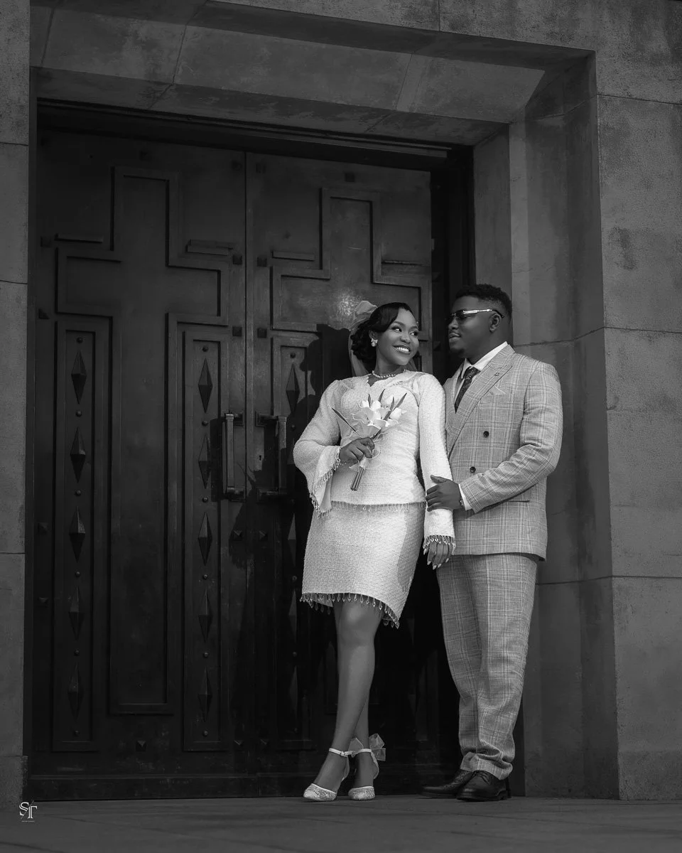 A smiling She and She with a bouquet of flowers are standing in front of a large door, possibly at a wedding or formal event.
