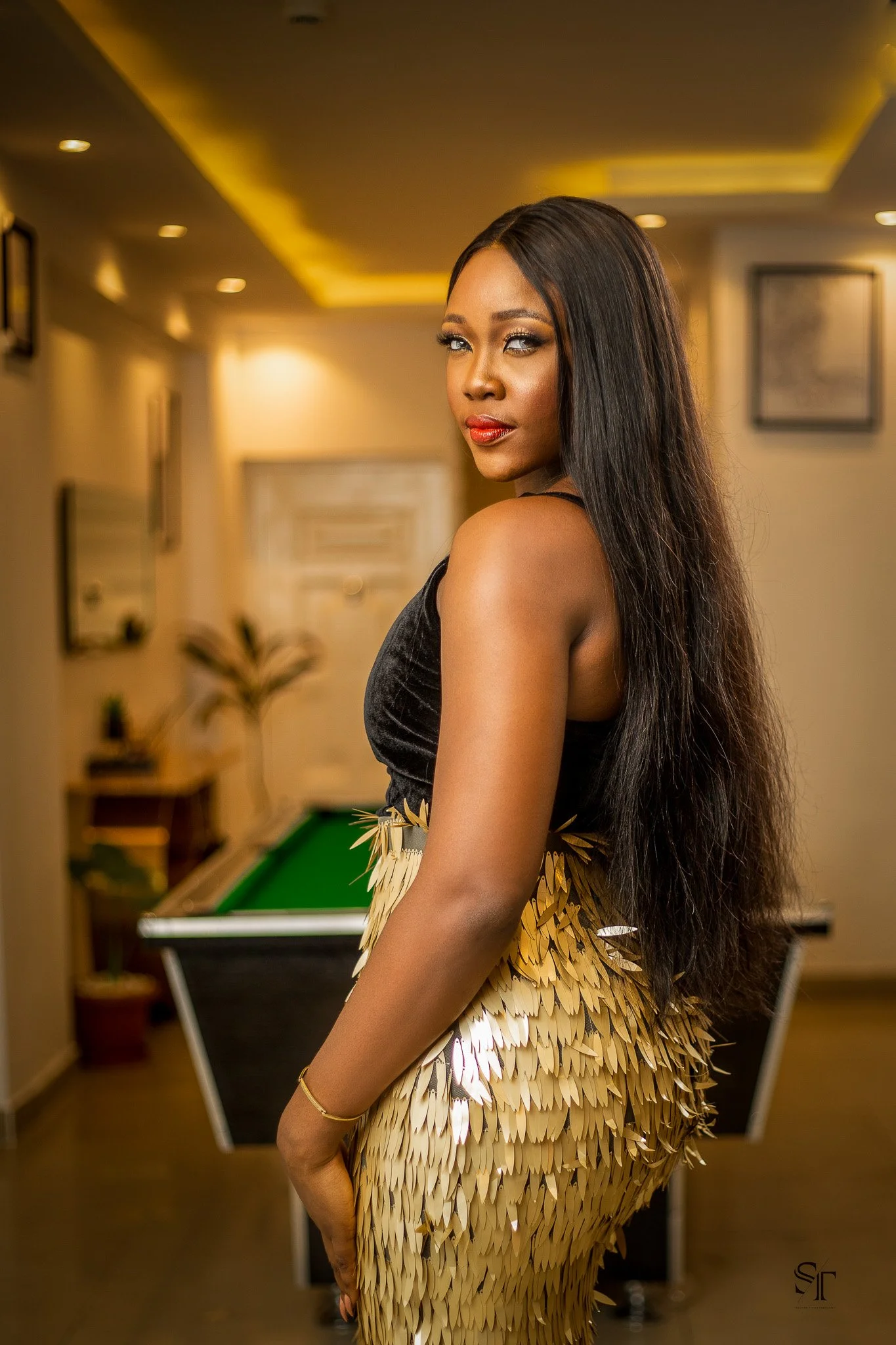 A woman with long dark hair and makeup, wearing a black sleeveless top and a gold sequined skirt, stands in a room with warm lighting and a pool table in the background.