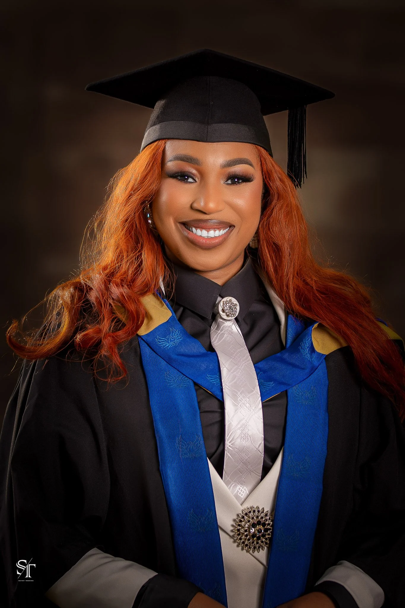 A woman in graduation attire wearing a black cap and gown with a blue and white stole, smiling at the camera. She has long, wavy red hair and is wearing earrings and a brooch.