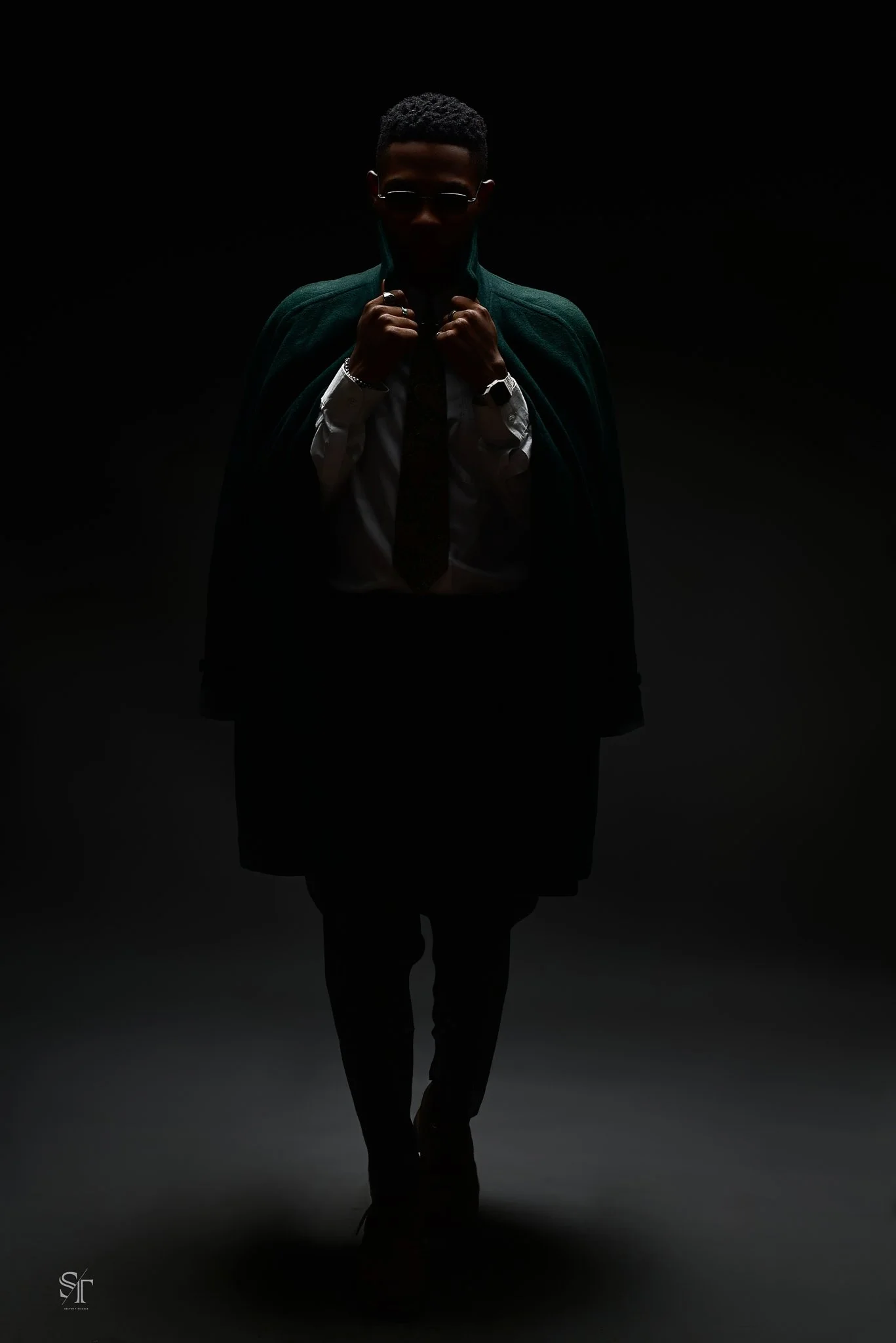 A person stands in a dark setting, wearing glasses, a white shirt, a tie, and a dark coat, with a green cape or coat draped over shoulders, and holding the collar of their shirt.