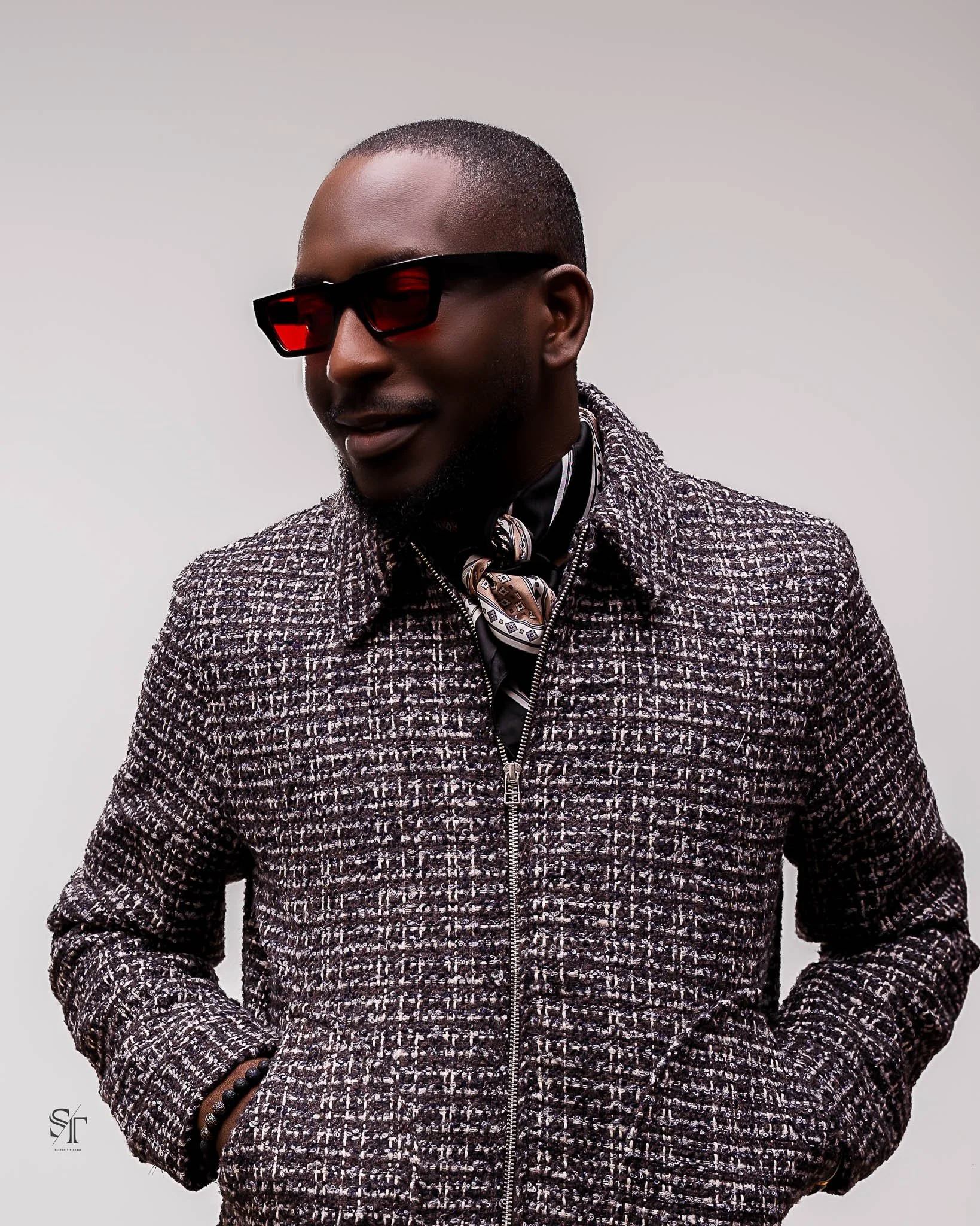 A man wearing red-tinted sunglasses, a patterned coat, and a scarf, standing against a plain background.