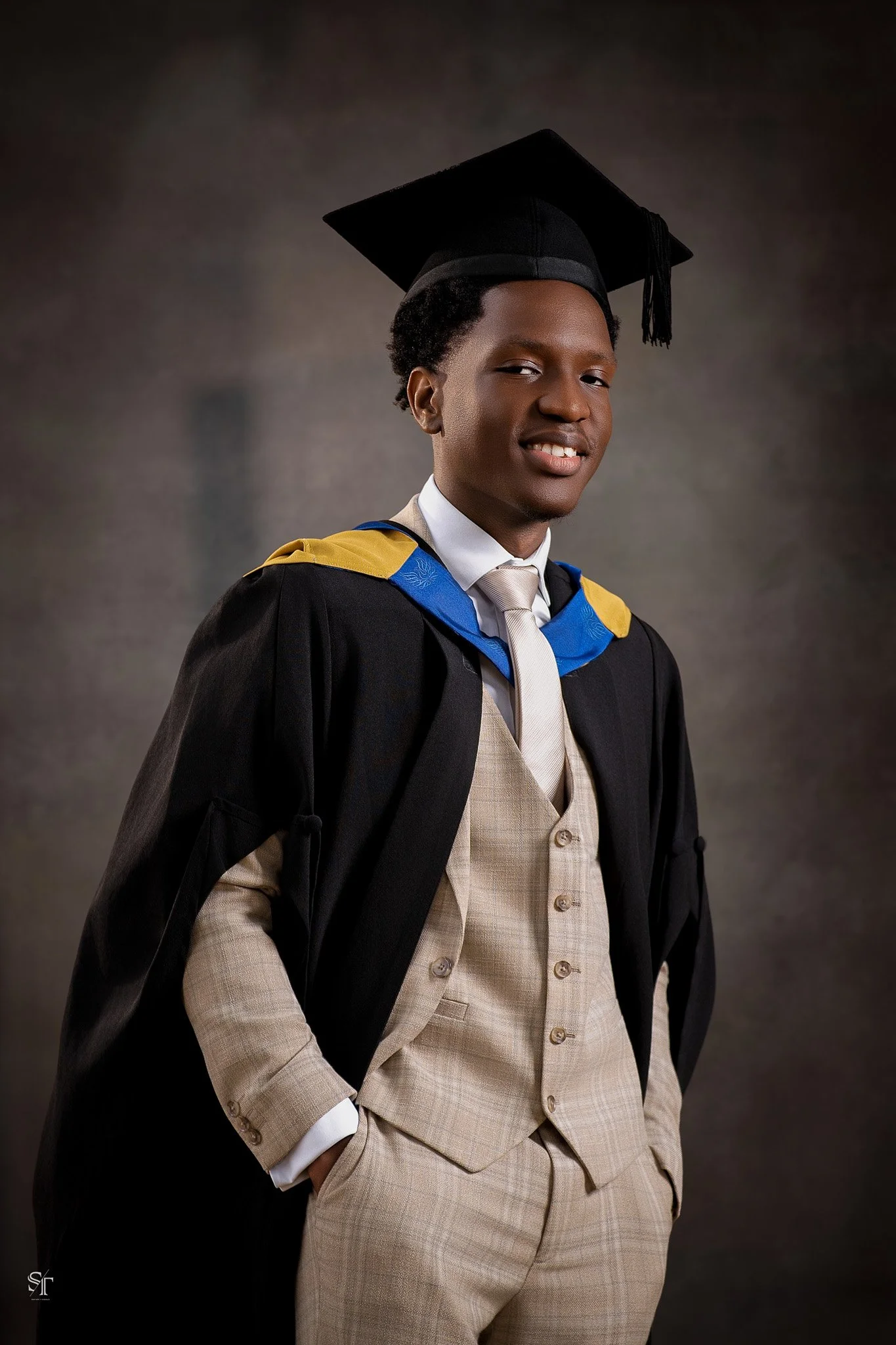 A young man in graduation attire, including a black cap and gown with blue and yellow accents, a white shirt, beige vest, and tie, posing with his left hand in his pocket against a plain dark background.