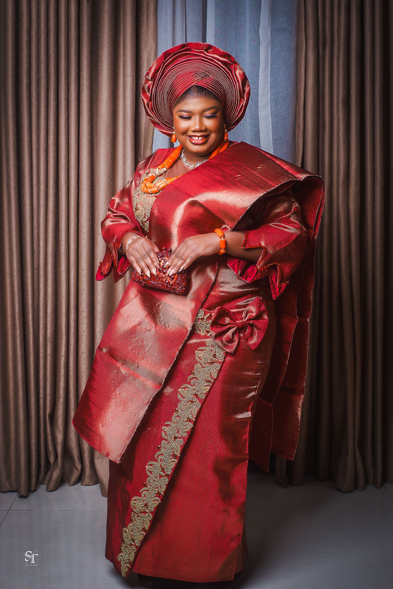 A woman dressed in traditional Nigerian attire, including a red and gold Ankara fabric, gele headwrap, and matching accessories, standing in front of a curtain.