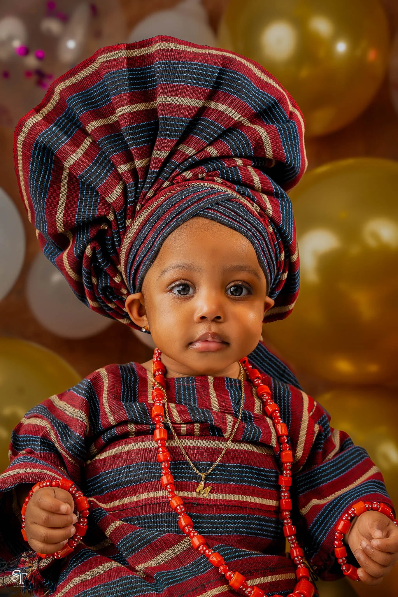 Young girl in traditional African attire with a large headwrap, red beaded jewelry, and a matching dress, standing in front of gold and white balloons.
