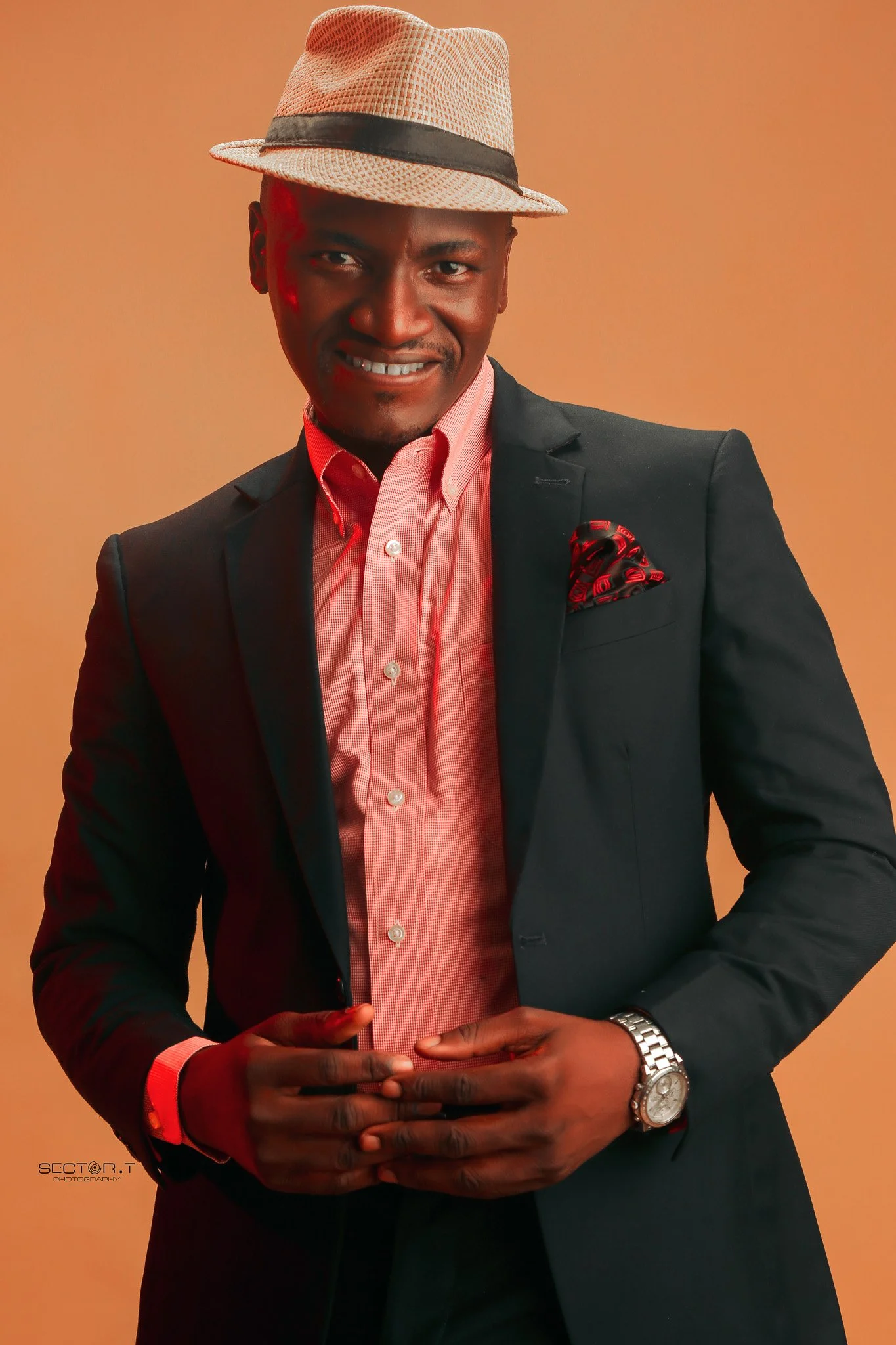 A man wearing a beige fedora hat, black blazer, pink button-up shirt, and a patterned pocket square, standing against a neutral background.