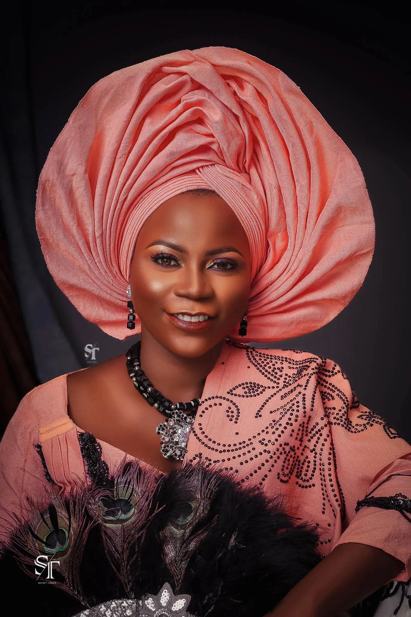 A woman wearing a coral pink traditional outfit with intricate black embroidery, a matching large gele headwrap, and black feathered accessories, posing for a portrait against a dark background.