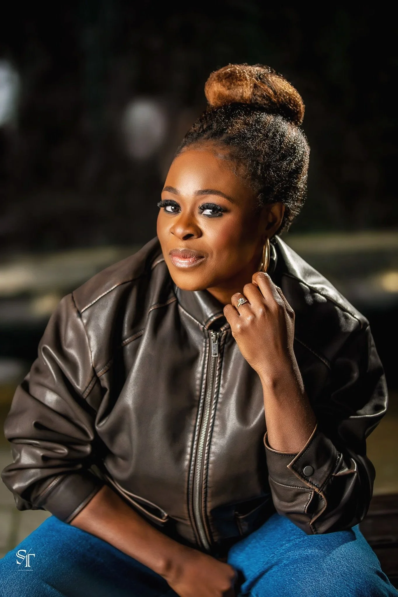 A woman with dark skin, wearing a leather jacket and hoop earrings, sitting outdoors at night with a confident expression.