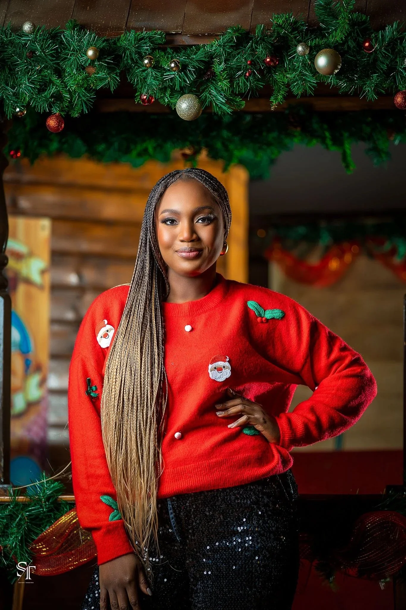 A woman wearing a red Christmas sweater with Santa and holly decorations, standing indoors in front of Christmas decorations and greenery.
