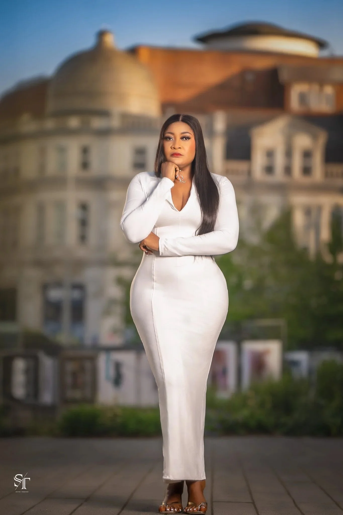 A woman with long black hair wearing a white, form-fitting dress and high heels, standing outdoors in front of a blurred, large, historic building.