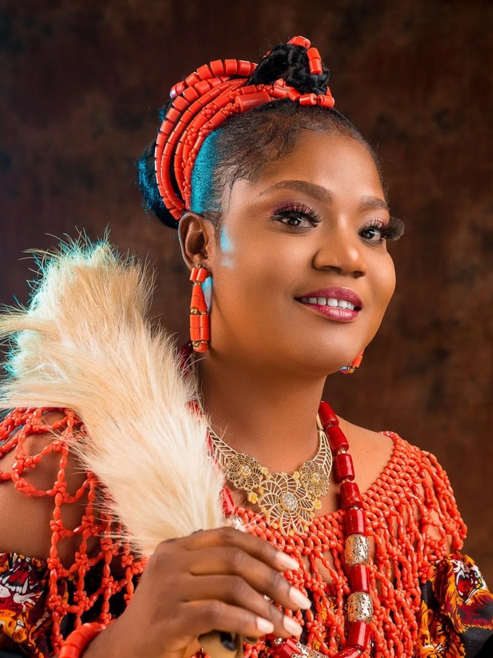 A woman dressed in traditional African attire with red bead jewelry, a red headwrap, and holding a white feather, standing against a dark, textured background.