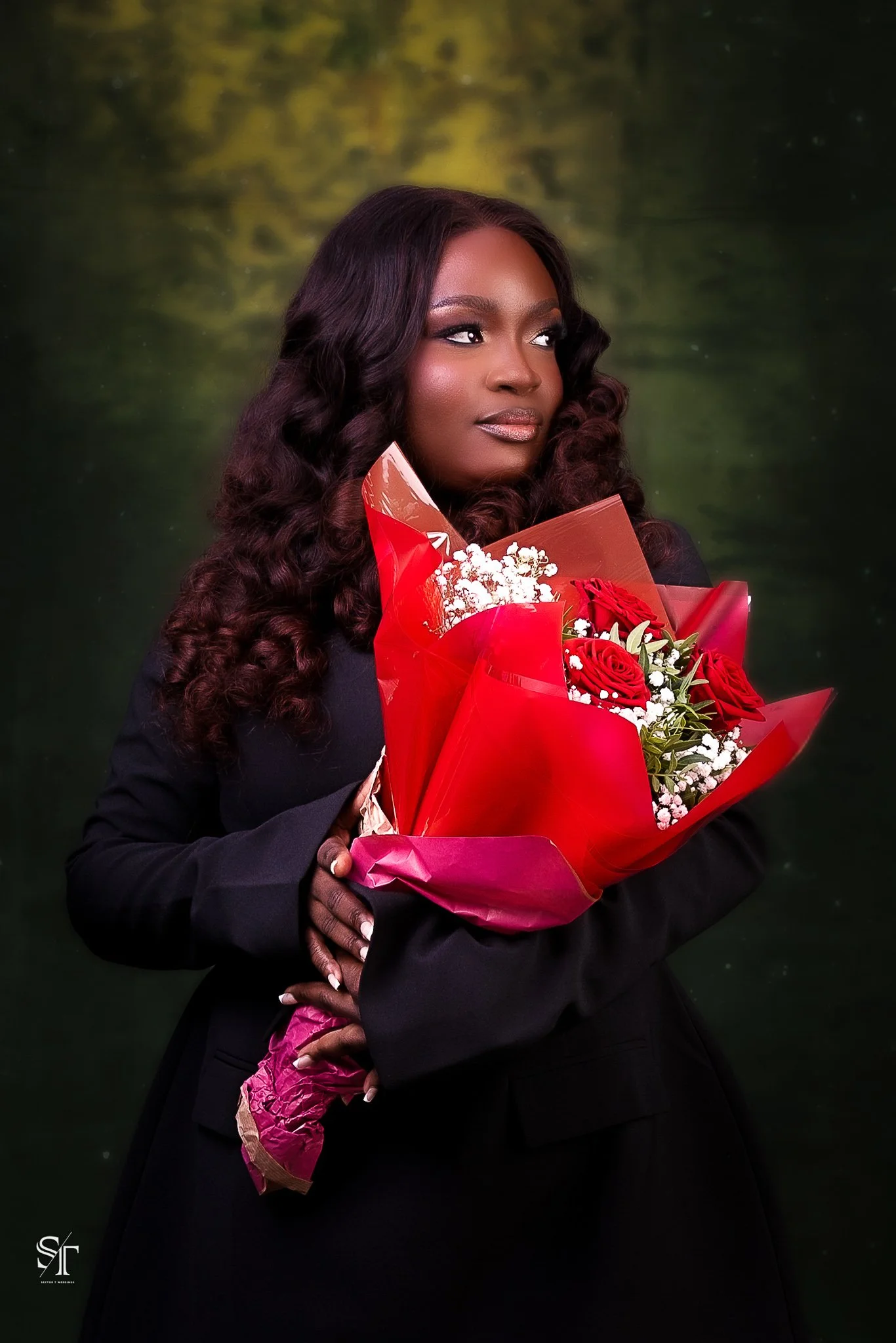 A woman with long, wavy hair holding a bouquet of red roses and white flowers, dressed in a black outfit, standing against a green and yellow blurred background.