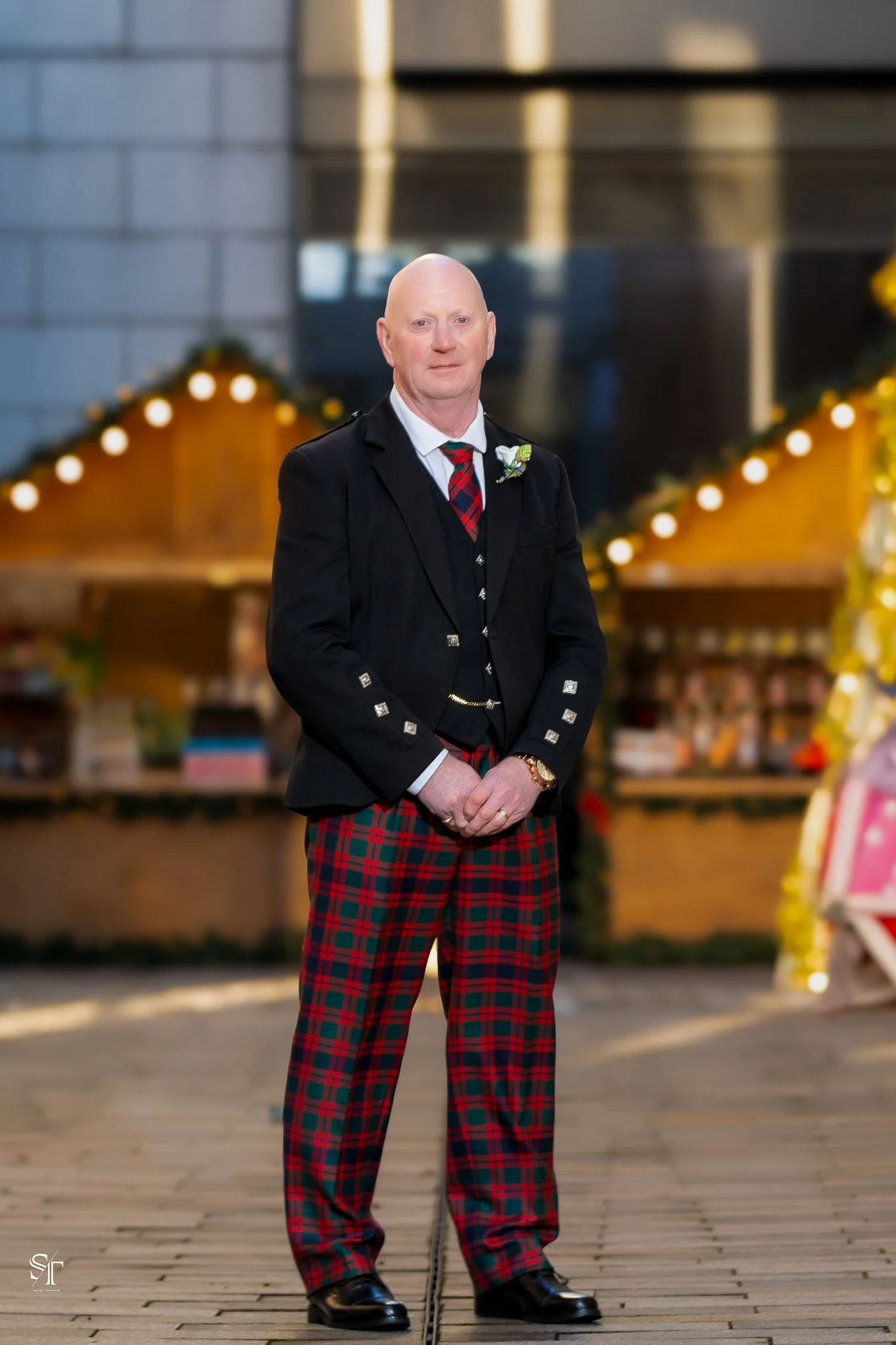 A man dressed in traditional Scottish attire, including a black blazer with silver buttons, a red and green plaid kilt, and a white shirt with a striped tie. He is standing outdoors on a wooden surface, with a blurred background featuring festive lig