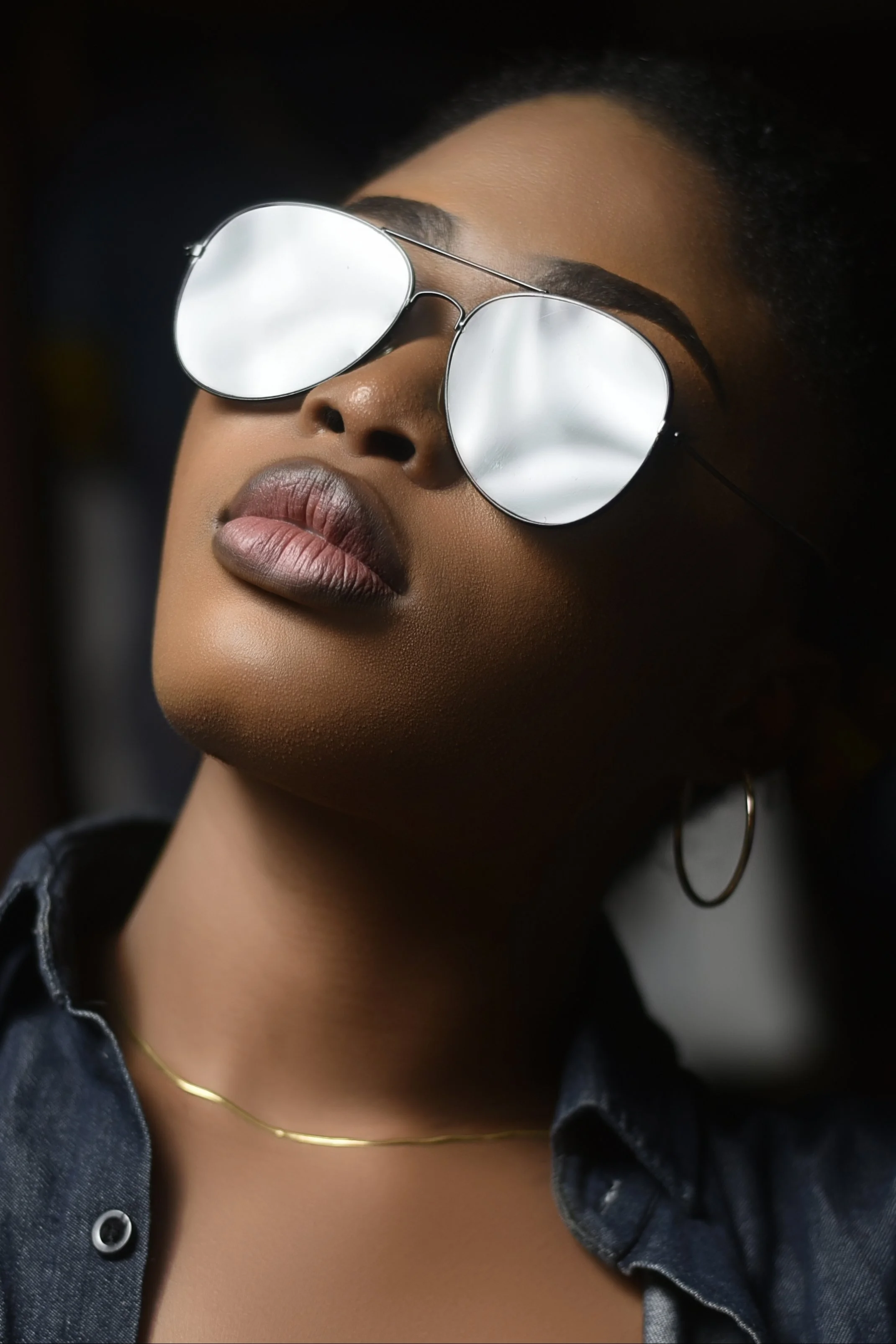 Close-up of a woman wearing reflective sunglasses, hoop earrings, a thin gold necklace, and a dark denim shirt with a collar.