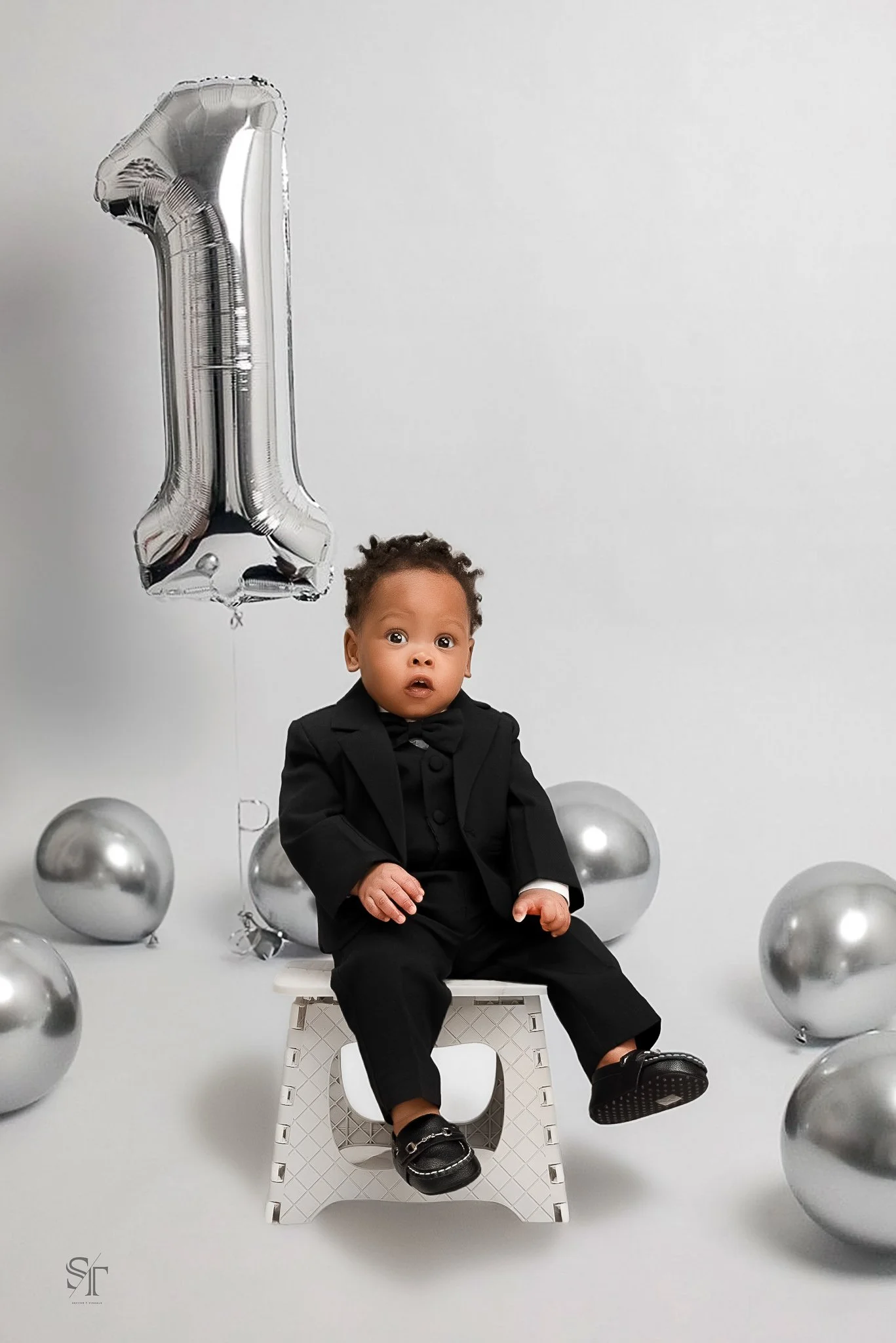 A baby dressed in a black suit, sitting on a small white chair, surrounded by silver balloons, with a large silver number one balloon behind him, celebrating his first birthday.