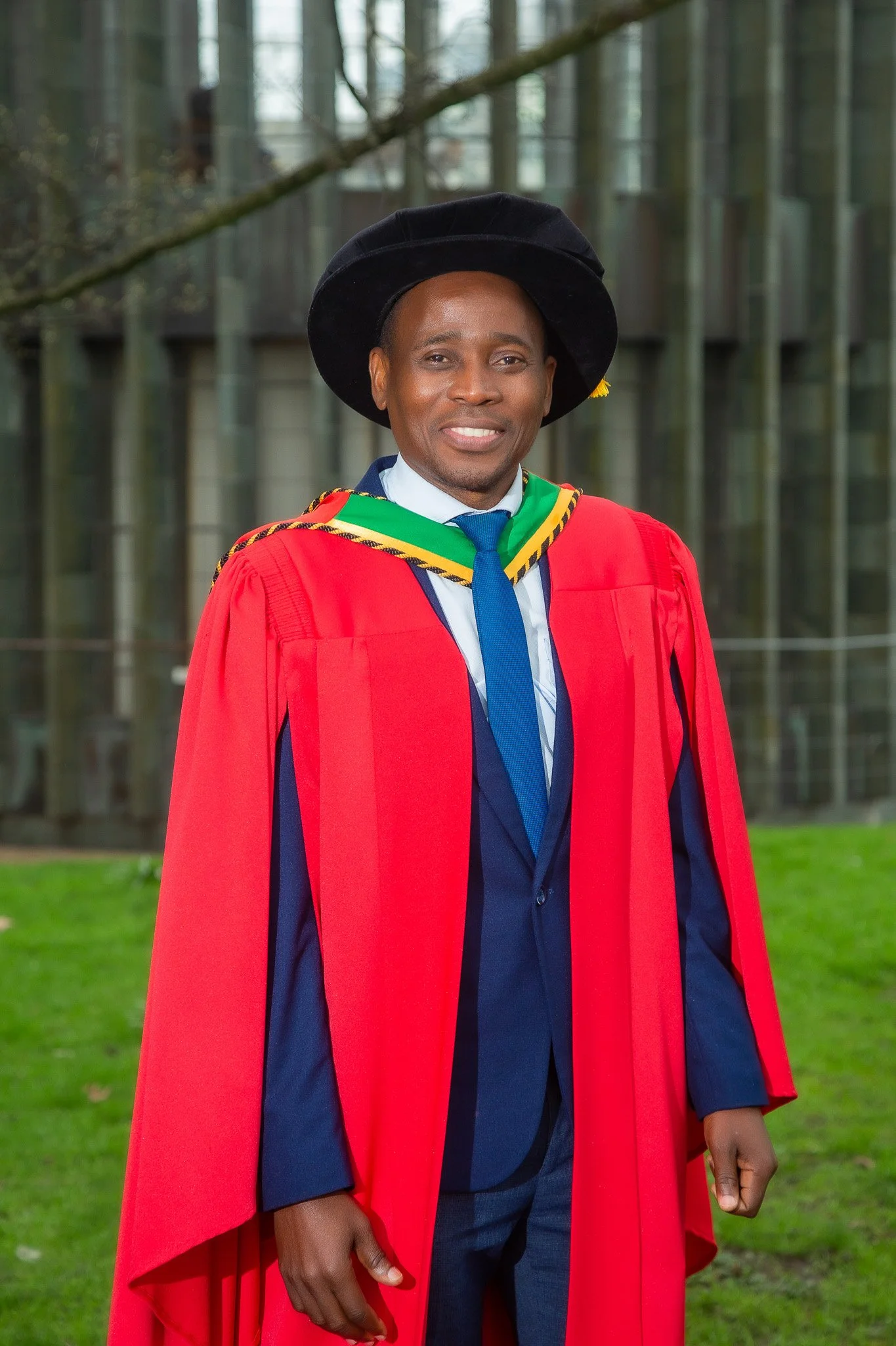 Man in graduation gown and cap standing outdoors with glass building in background.