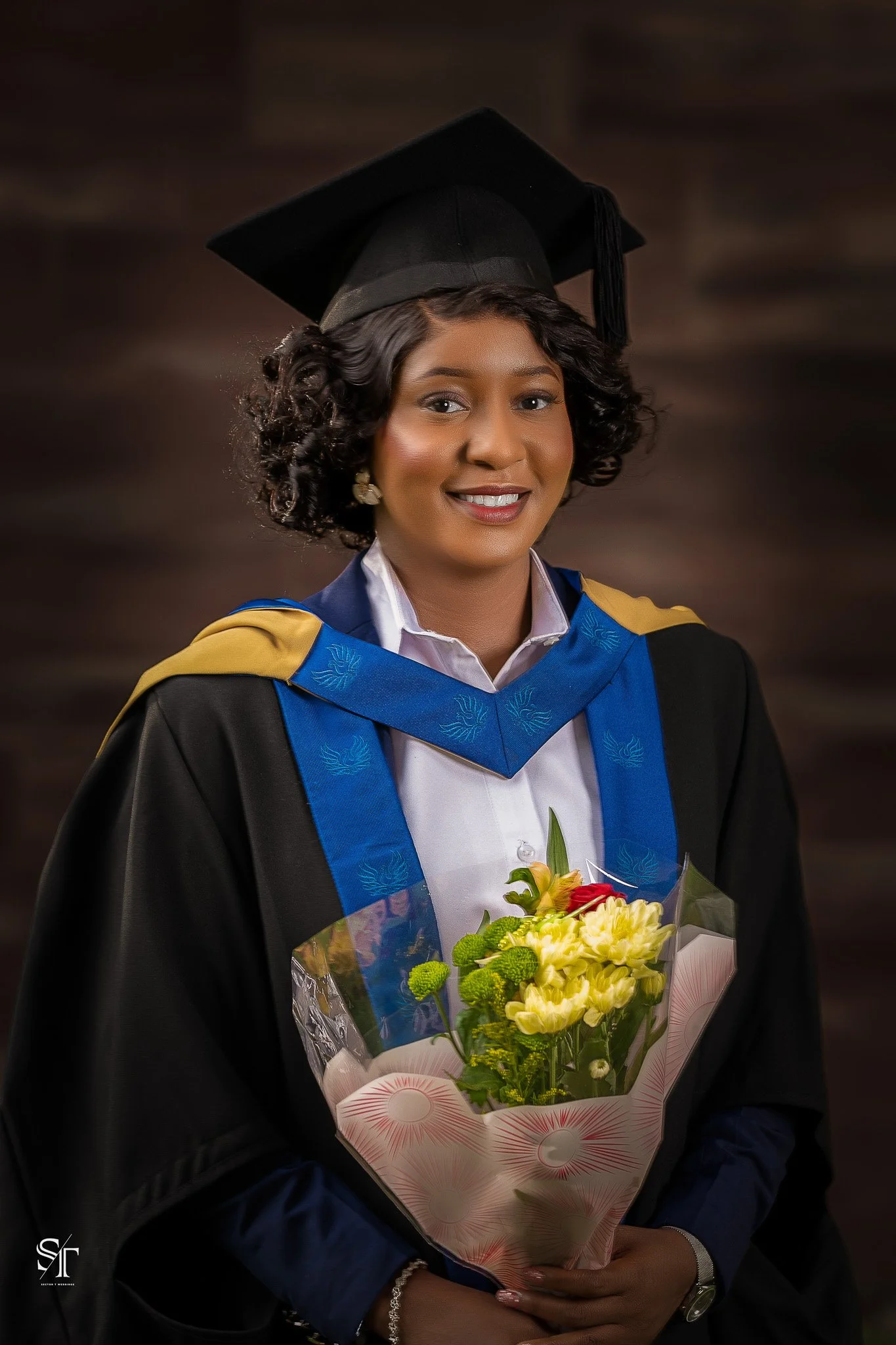 A young woman in graduation cap and gown holding a bouquet of flowers, smiling at the camera.