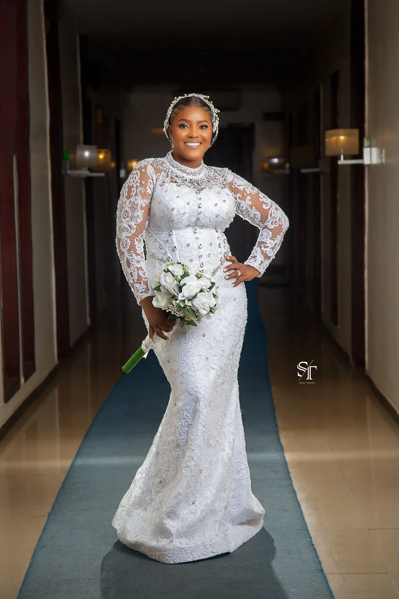 Smiling bride in a white lace wedding gown with long sleeves, holding a bouquet of white flowers, standing in a dimly lit hallway.