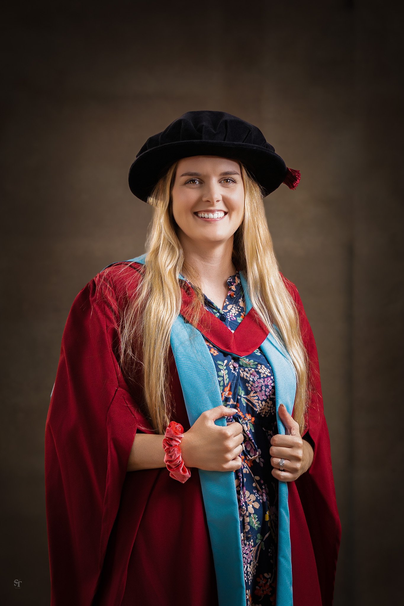 A woman smiling and wearing a graduation cap and gown, standing against a brown background.