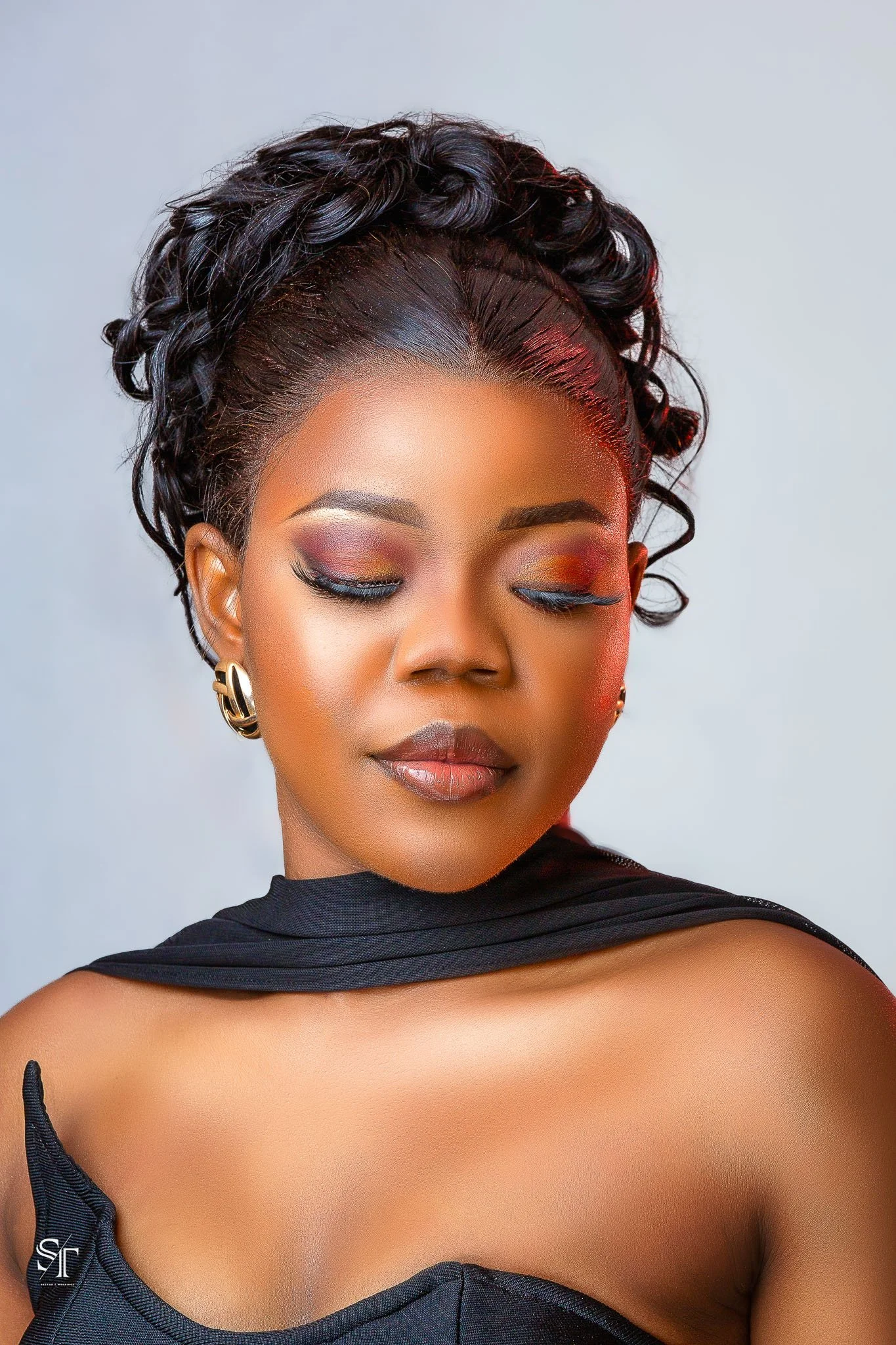 A woman with dark, curly hair styled in an elegant updo, wearing makeup with colorful eyeshadow, and gold hoop earrings. She has her eyes closed and is dressed in a black strapless top with a matching black wrap around her shoulders.