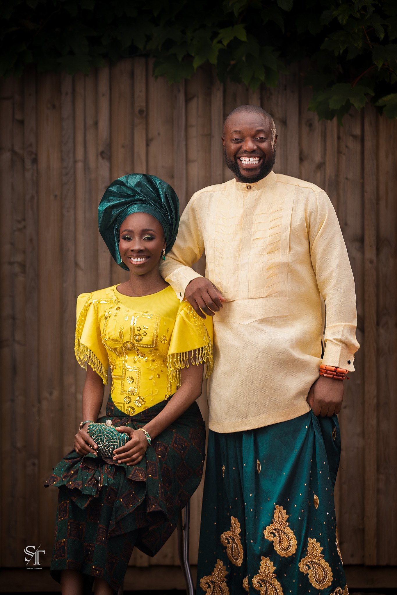 A smiling man and woman dressed in traditional African attire standing in front of a wooden fence and green foliage.