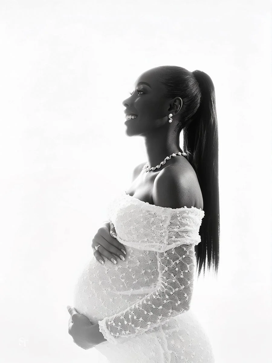Black and white photo of a pregnant woman smiling, wearing lace dress and jewelry, with her hands on her belly.