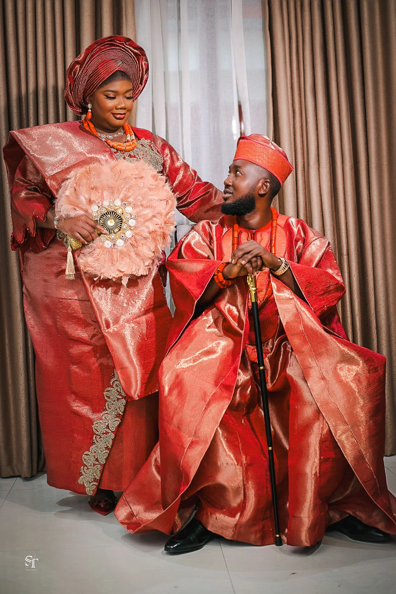 A couple dressed in traditional Nigerian attire, the woman standing and the man sitting in a wheelchair, both wearing red and gold outfits, with the woman holding a decorative fan.