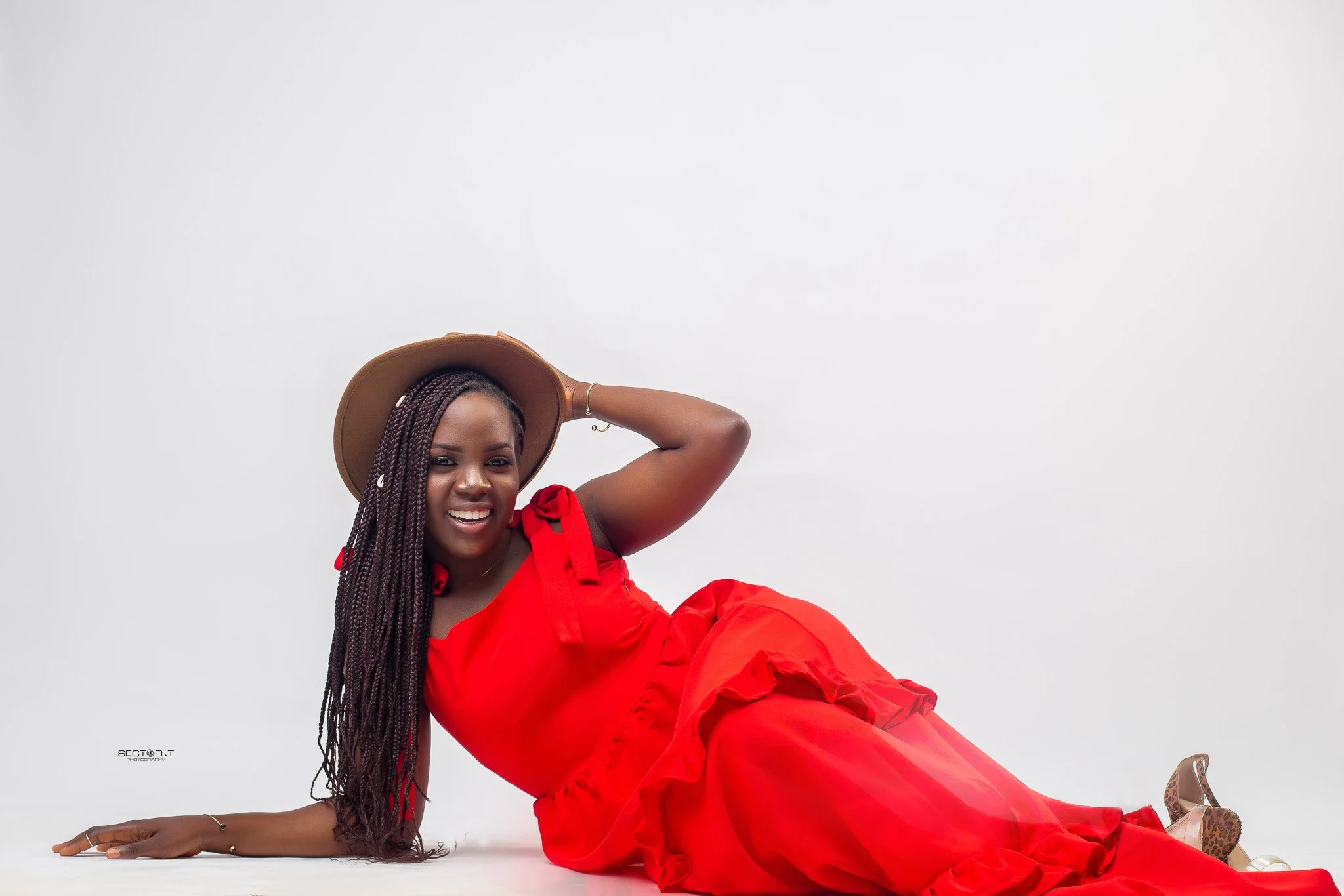 A woman in a red dress and a wide-brimmed hat, with long braided hair, lying on the floor and smiling at the camera against a plain white background.