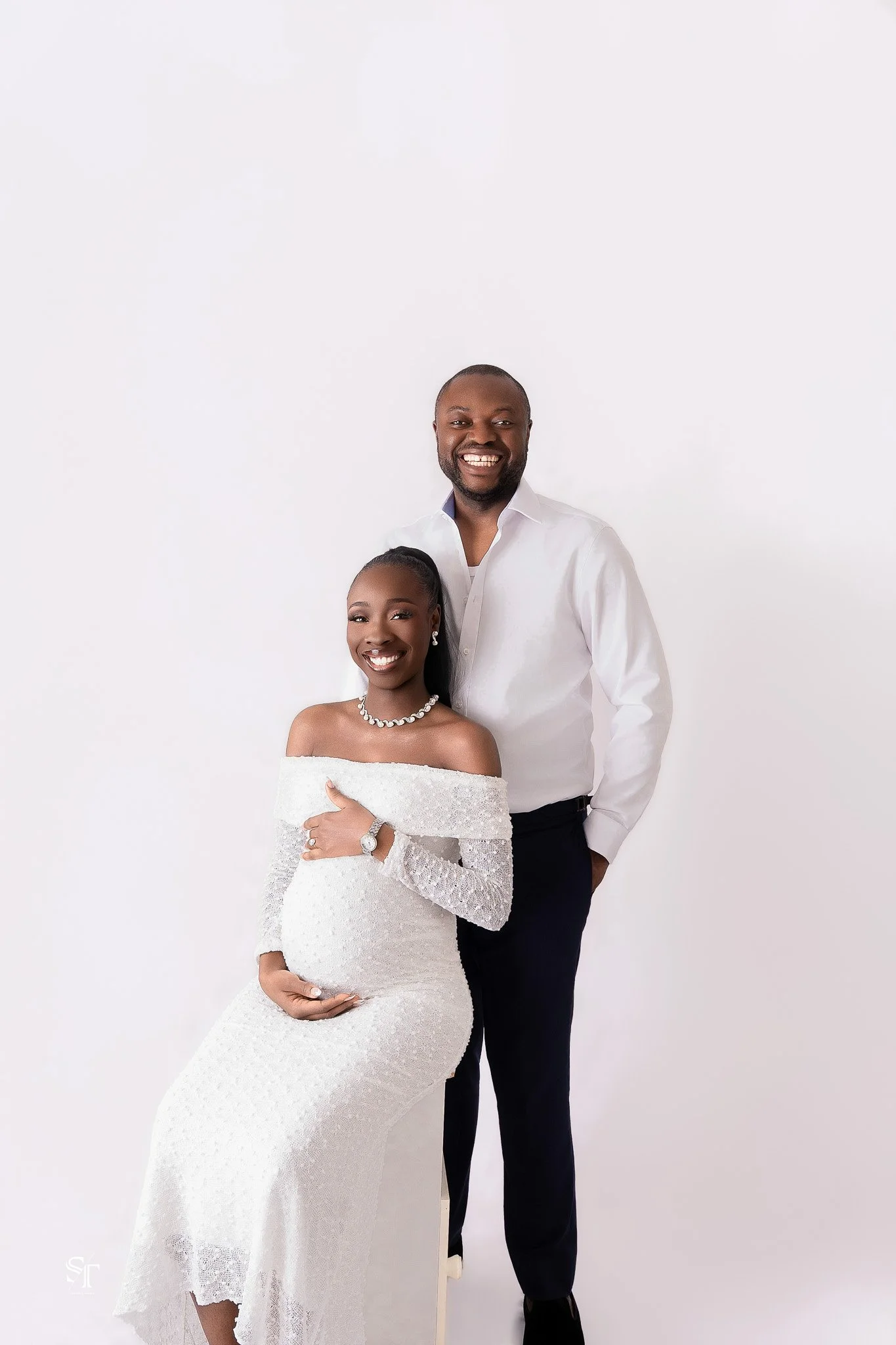 A smiling woman in a white off-shoulder wedding dress with jewelry, and a smiling man in a white shirt and black trousers, standing against a plain white background.