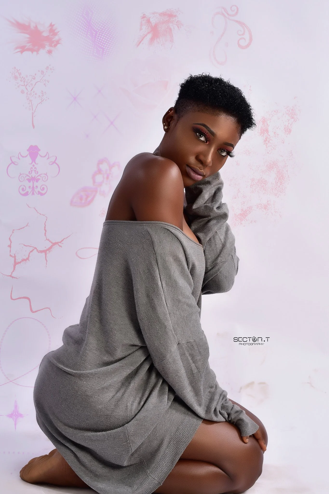 A young woman with dark skin and short curly hair kneels on the floor against a light background with pink abstract designs. She wears a loose gray sweater off her shoulder and looks directly at the camera.