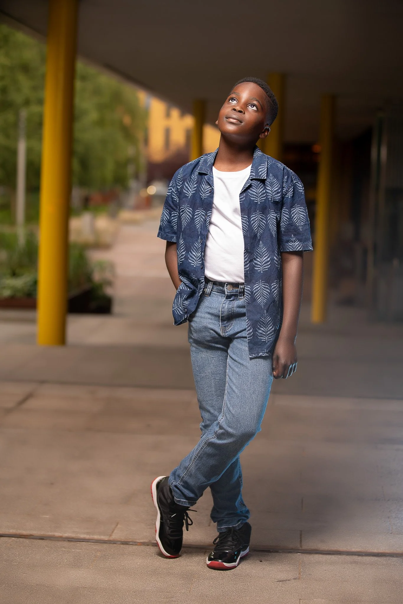 A young boy standing outdoors on a sidewalk in front of a building with yellow support columns, wearing a patterned blue short-sleeve shirt, white t-shirt, jeans, and black sneakers, looking upwards.