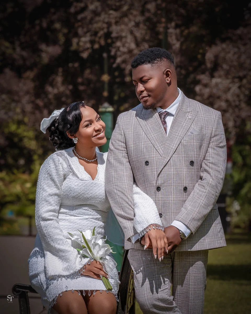 A couple dressed in wedding attire sharing an intimate moment outdoors in front of trees. The woman is wearing a white wedding dress, a pearl necklace, earrings, and has a bouquet of calla lilies. The man is in a gray checkered suit with a white shir