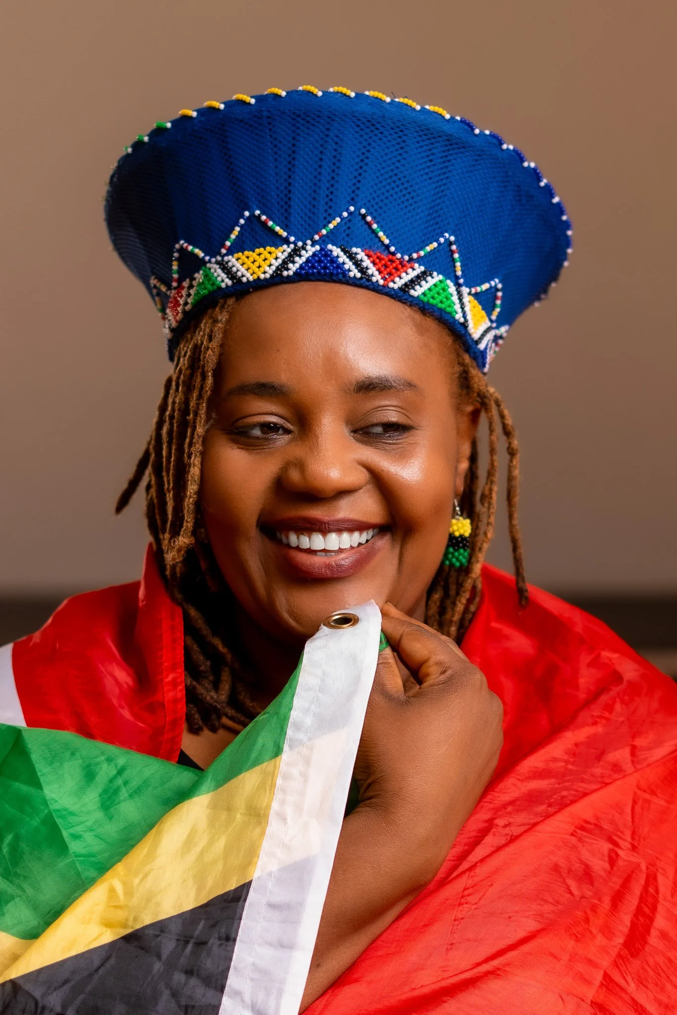 Smiling woman wearing a blue beaded crown, earrings resembling the Tanzanian flag, and holding a Tanzanian flag, draped in a red fabric.