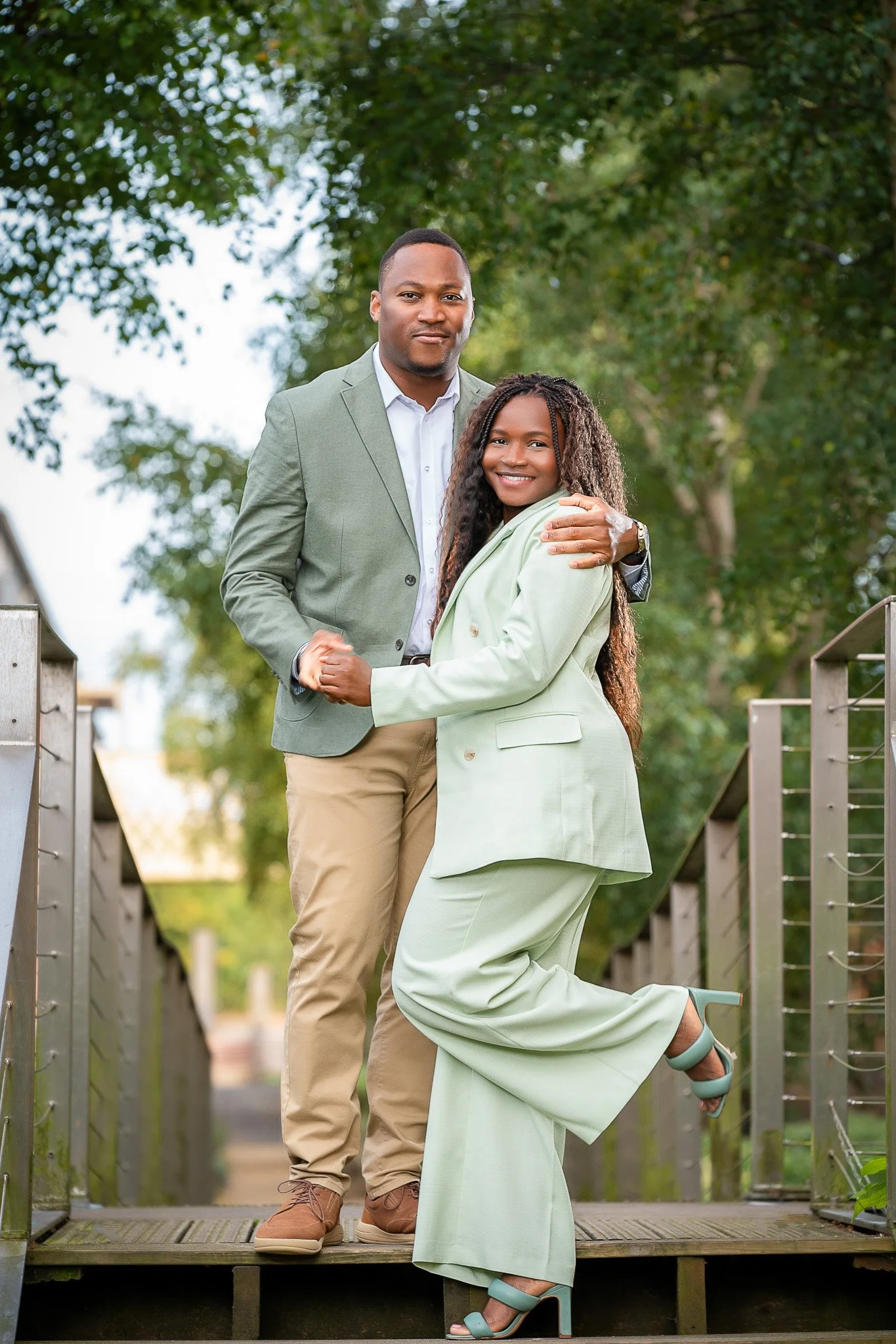 A man and a woman posing together outdoors on a bridge, with greenery in the background. The man is wearing a green blazer, white shirt, and khaki pants. The woman is wearing a light green suit and teal high heels. They are smiling and appear happy.