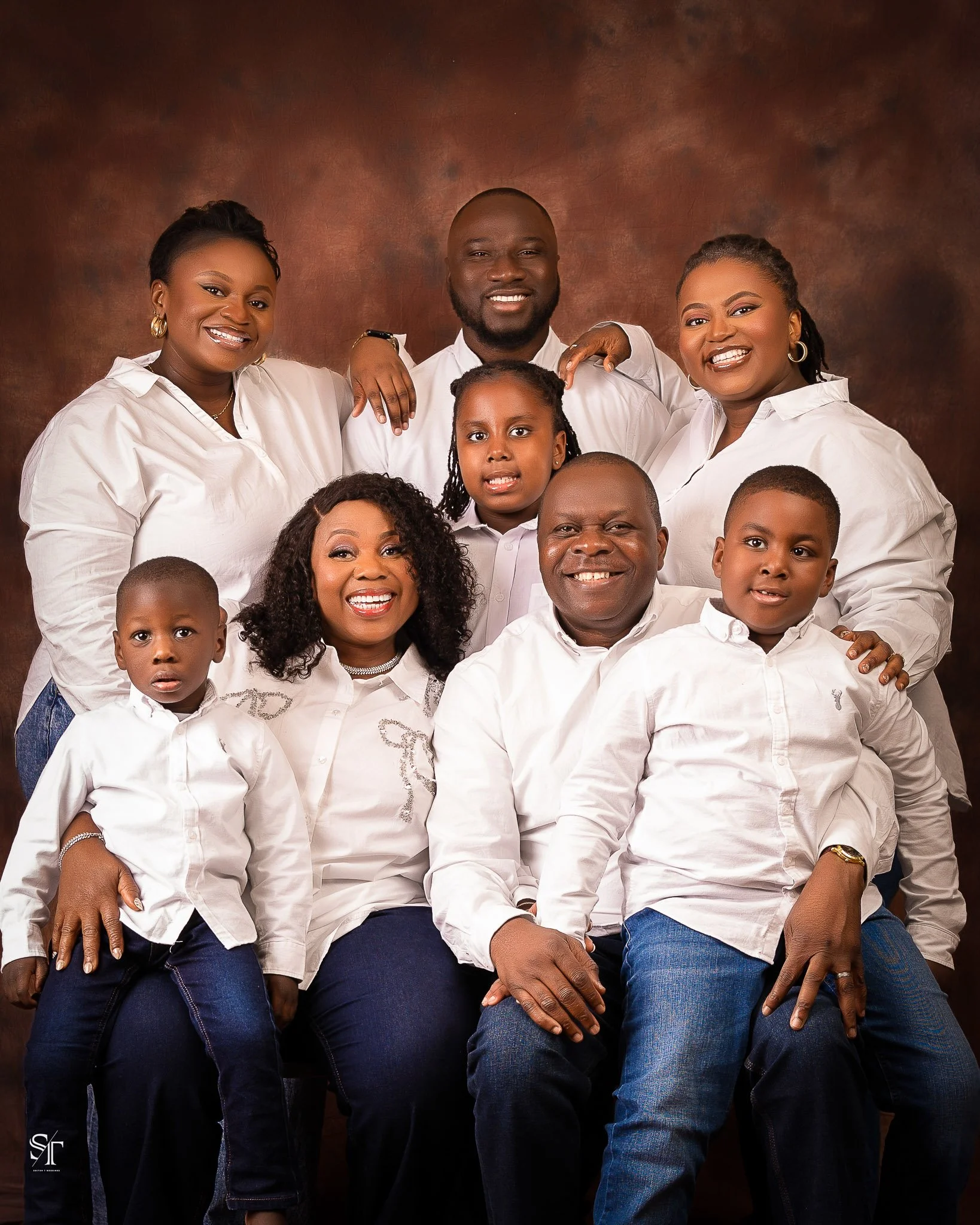 A large family portrait featuring nine people, including adults and children, all dressed in white shirts and dark jeans, posing against a brown background.