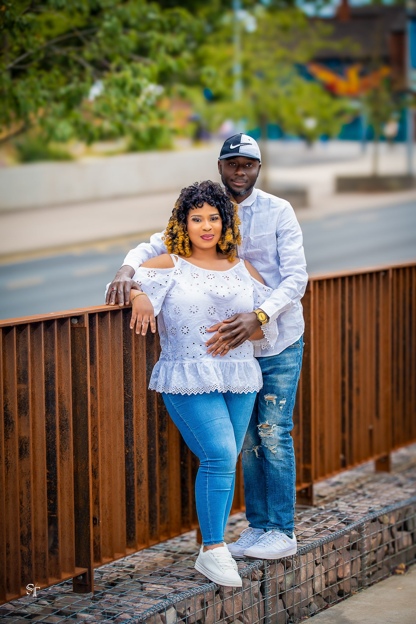 A couple standing on a bridge outdoors, with the man standing behind the woman, both leaning against a wooden railing. The woman has curly hair with blonde tips, wearing a white eyelet blouse and blue jeans. The man has short hair, wearing a white sh