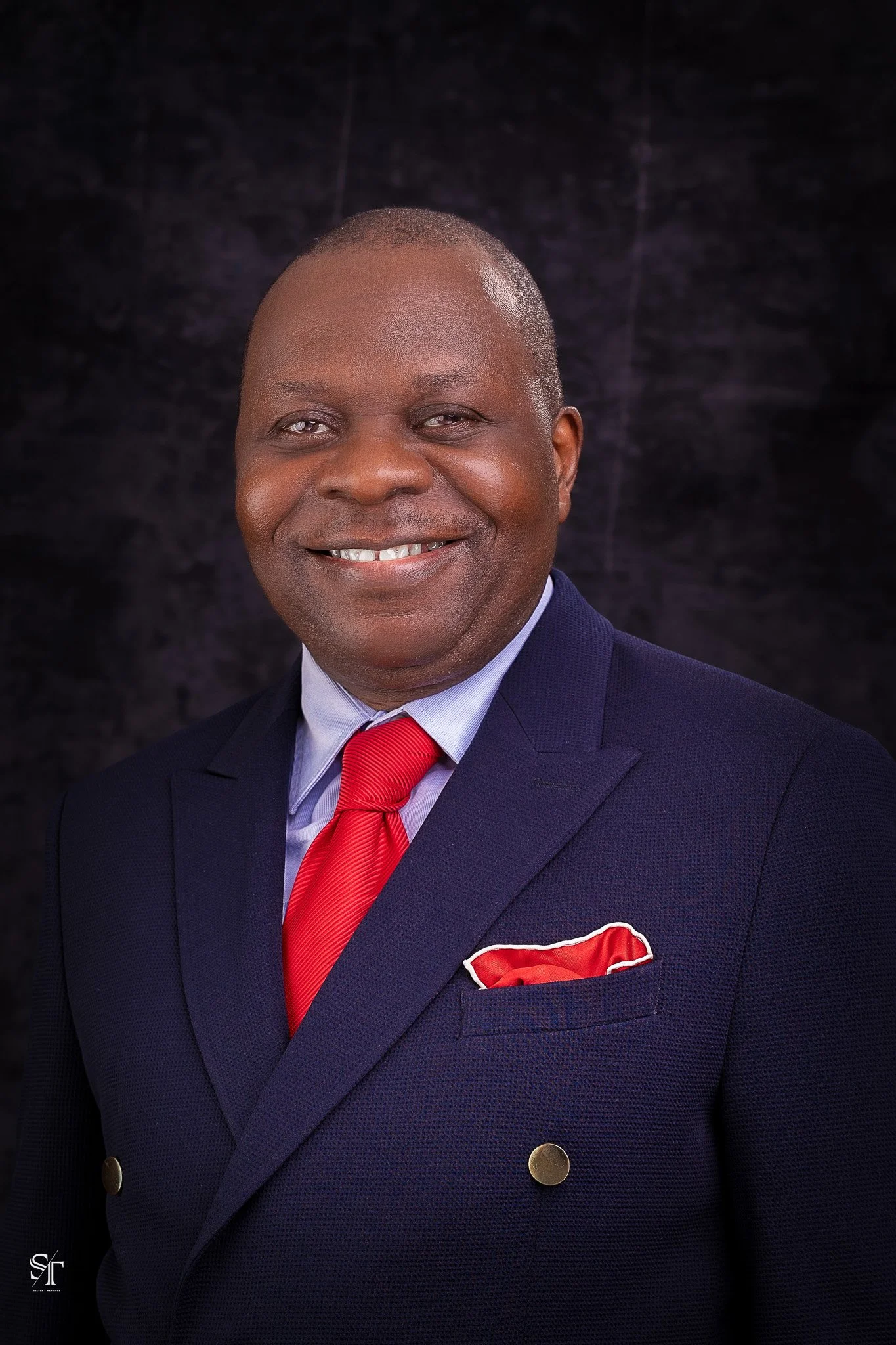 A smiling African American man wearing a dark navy suit with golden buttons, a light blue shirt, a red tie, and a matching red pocket square. He is standing against a dark background.