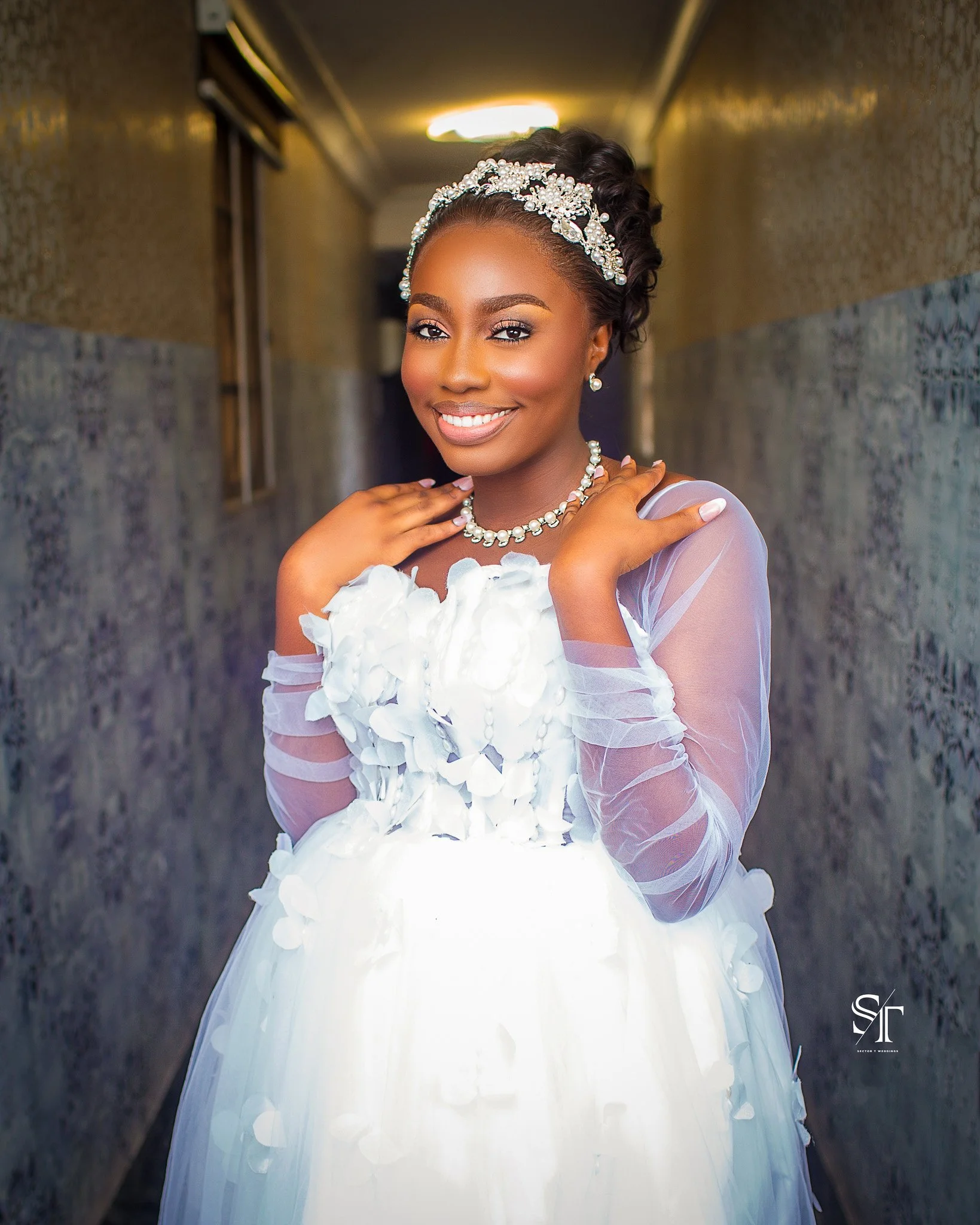 A bride smiling in a wedding dress with floral details and sheer long sleeves, wearing pearl jewelry and a tiara, standing in a corridor.