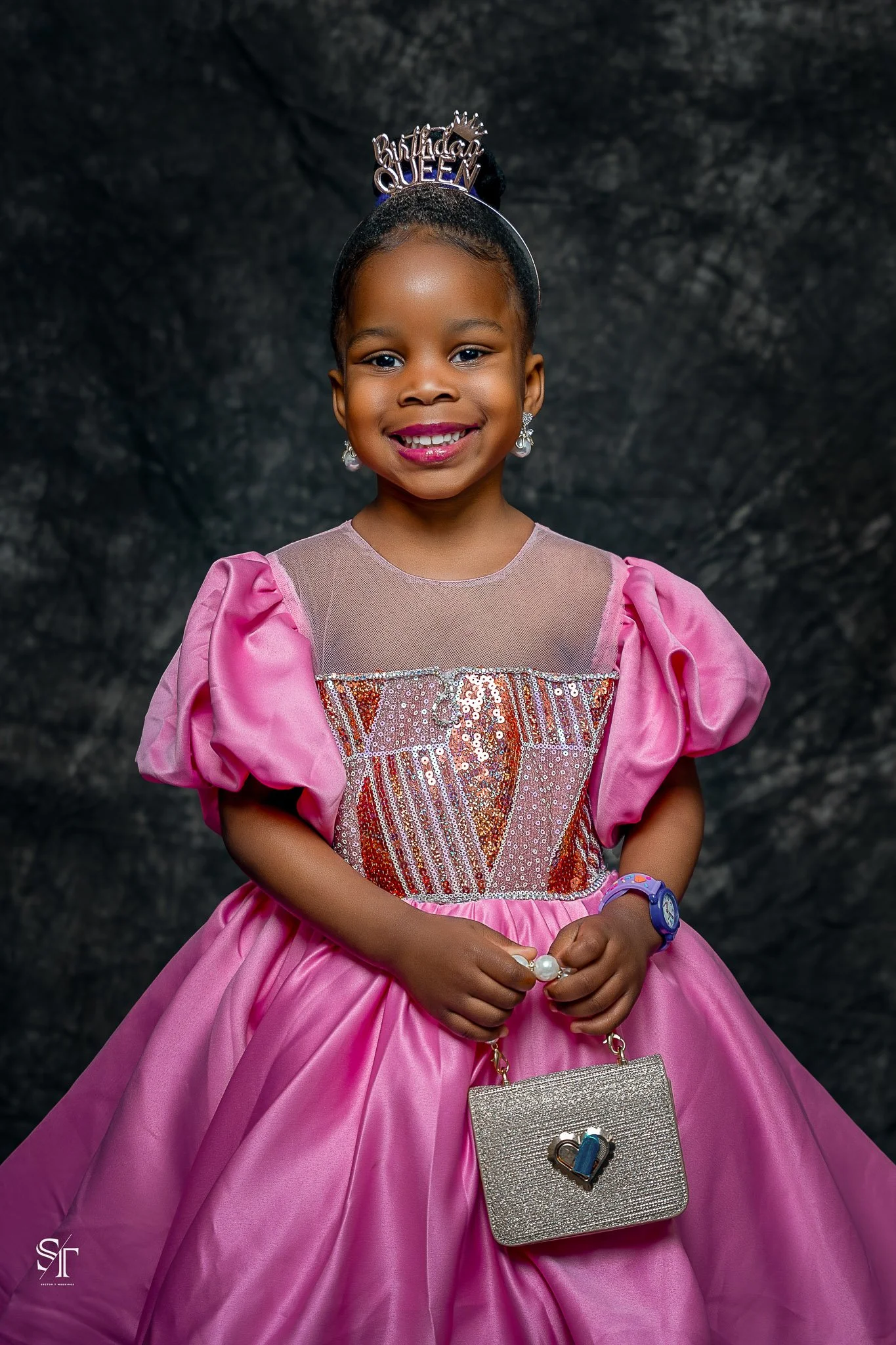 Young girl smiling in a pink princess dress with a "Birthday Queen" tiara and holding a small silver purse.