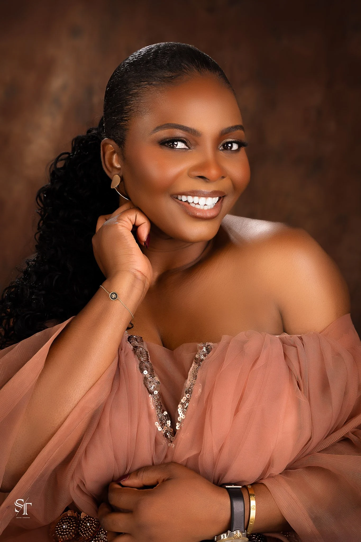 A woman with dark, curly hair styled in a ponytail, smiling and looking directly at the camera. She is wearing earrings, a delicate bracelet, and a watch with bracelets. Her off-shoulder dress is made of soft, pink fabric with sequins along the neckl