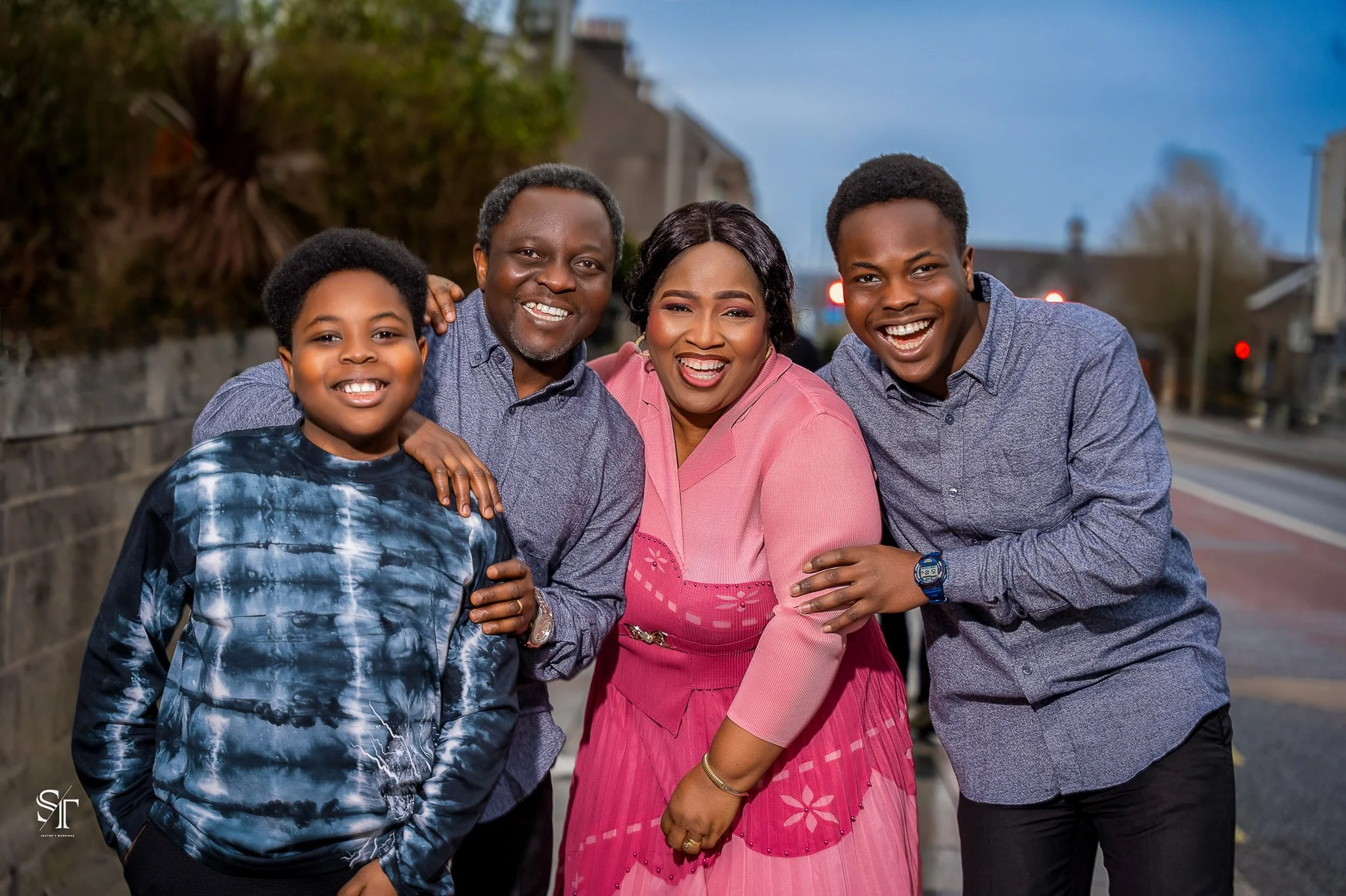 A happy multi-generational family of four standing outdoors on a city street at dusk, smiling and embracing each other.