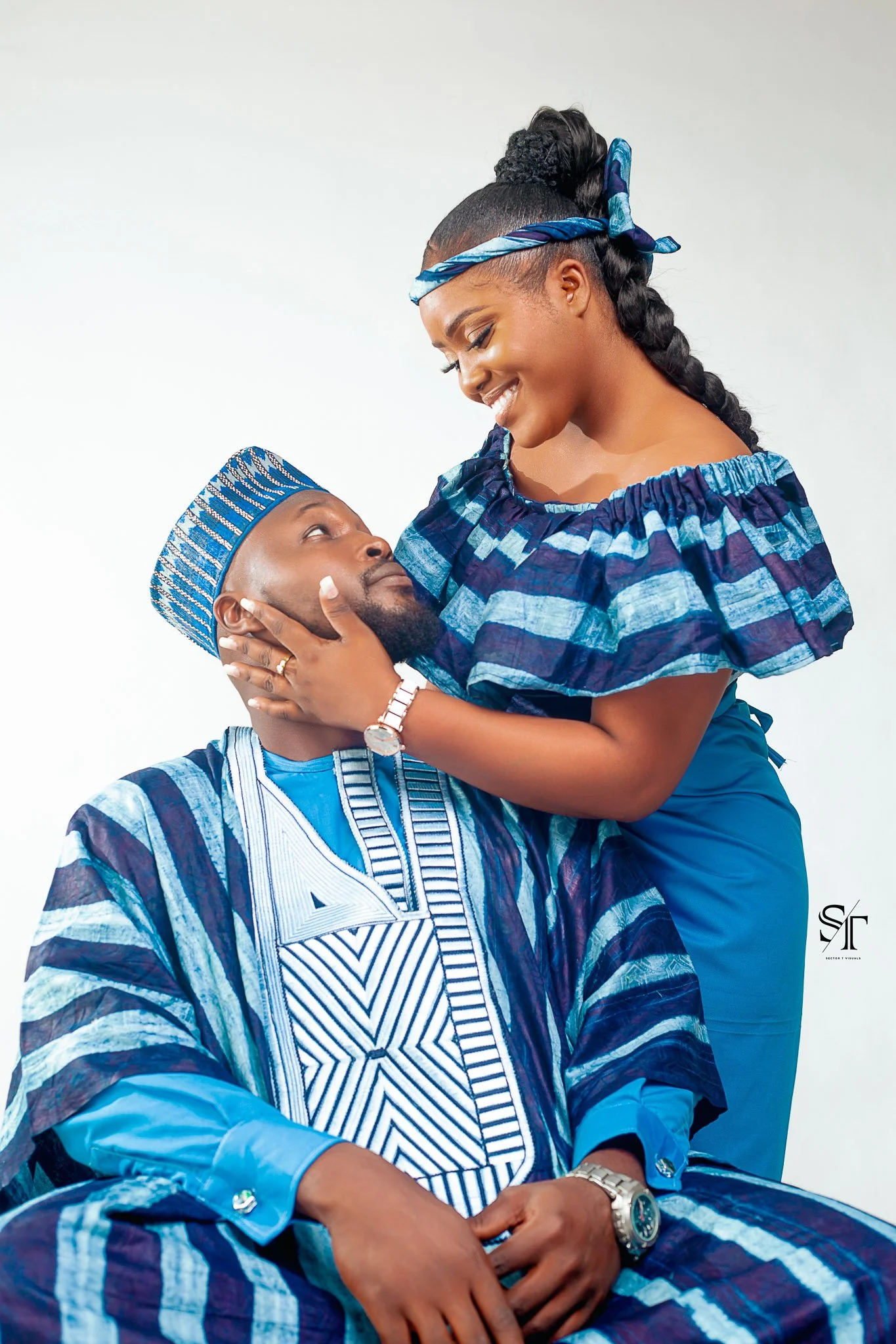 A woman and man dressed in traditional Nigerian attire, with the woman standing and gently holding the man's face as they look at each other affectionately.